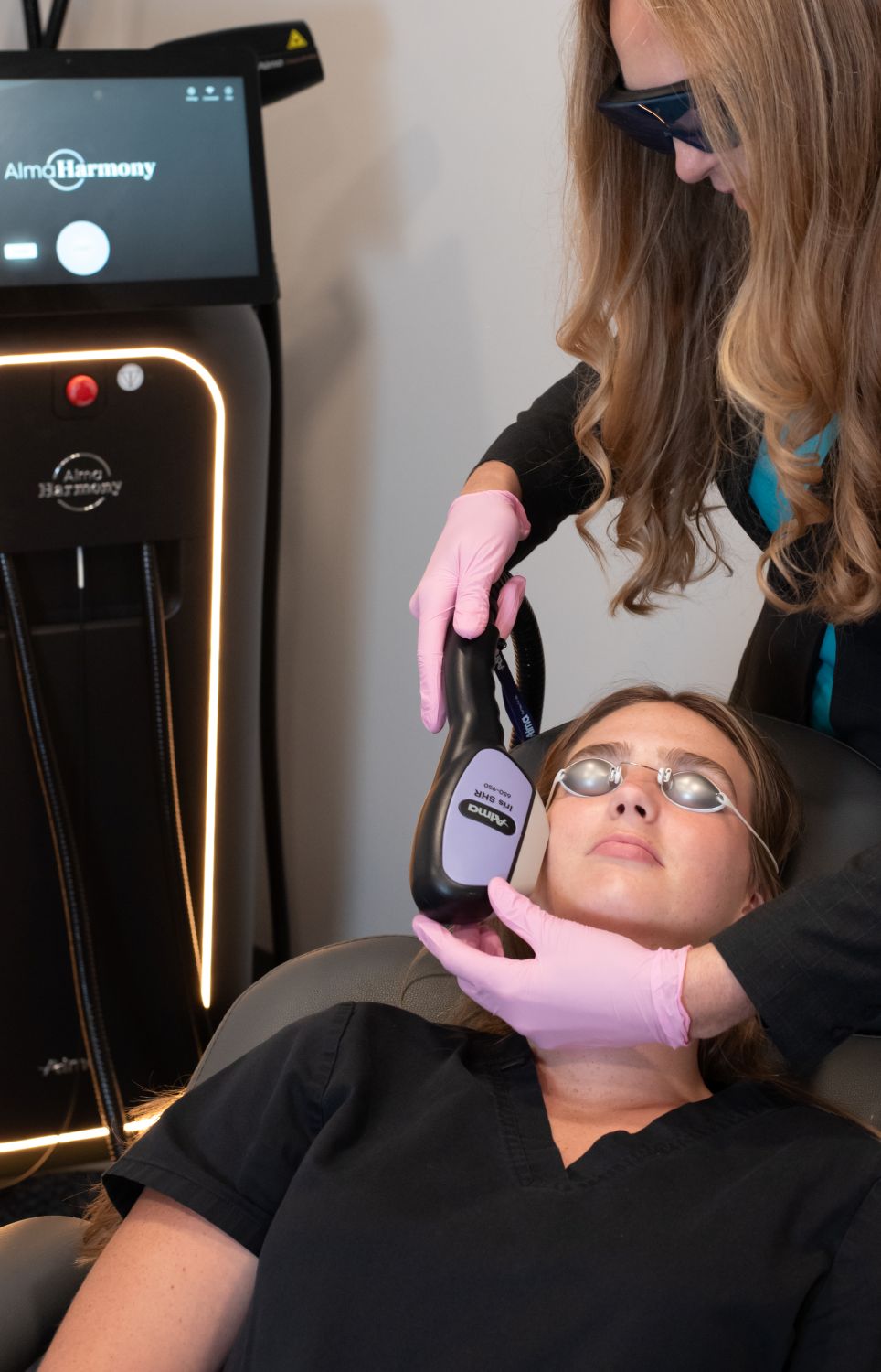 A woman with glasses lying down during a medical or cosmetic procedure, with a technician using equipment on her face.