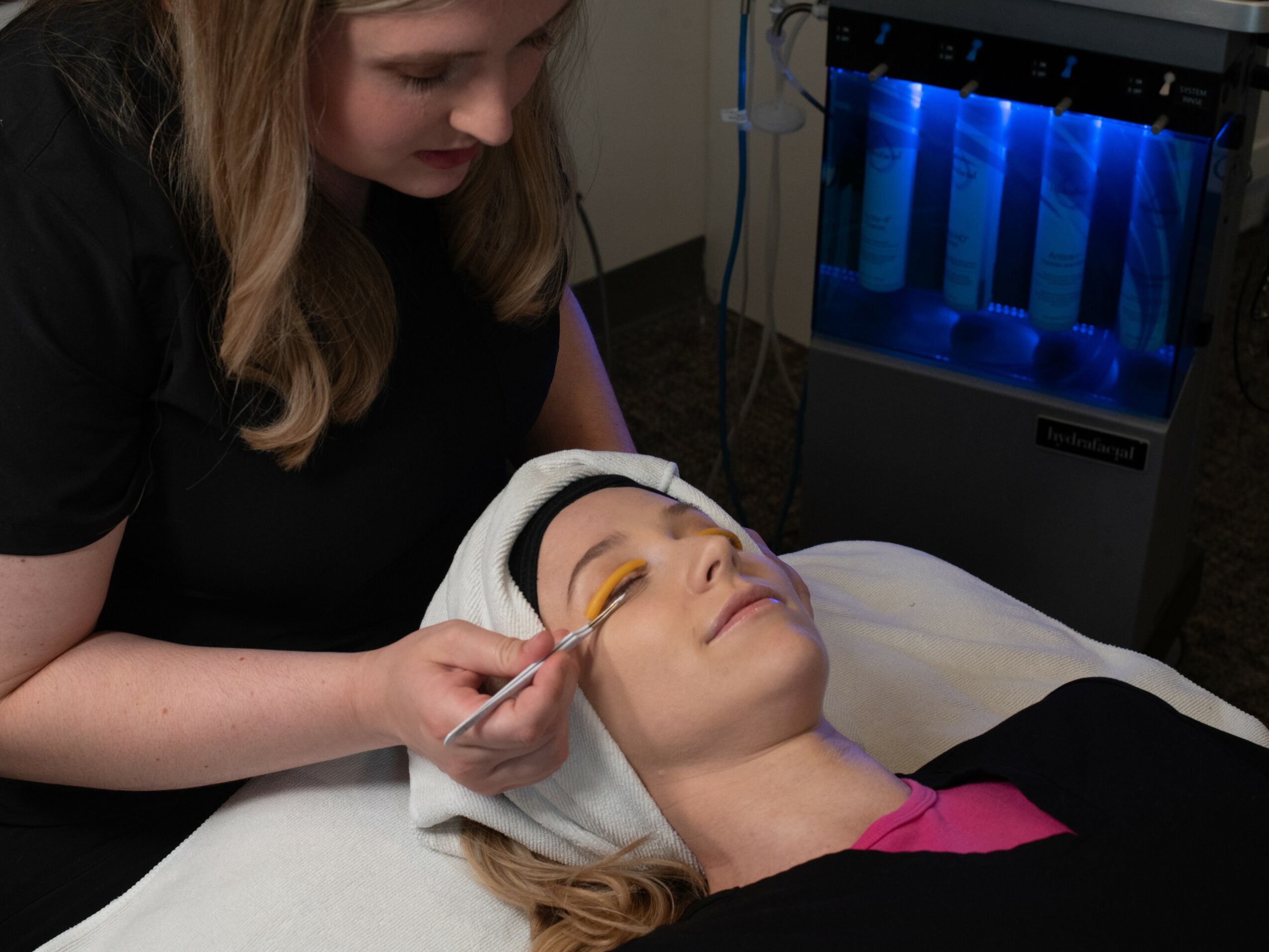 A woman applies makeup to a woman lying on a bed with a towel wrapped around her head, in a room with a blue-lit machine.