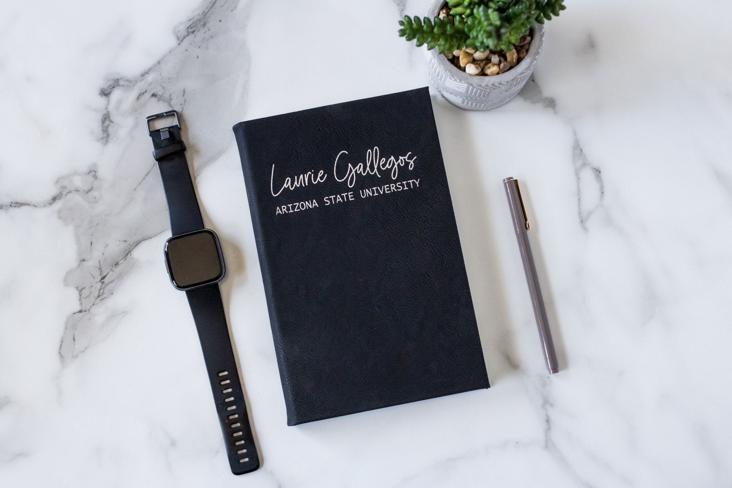 Black notebook with white text, a smartwatch, a pen, and a potted plant on a white marble surface.