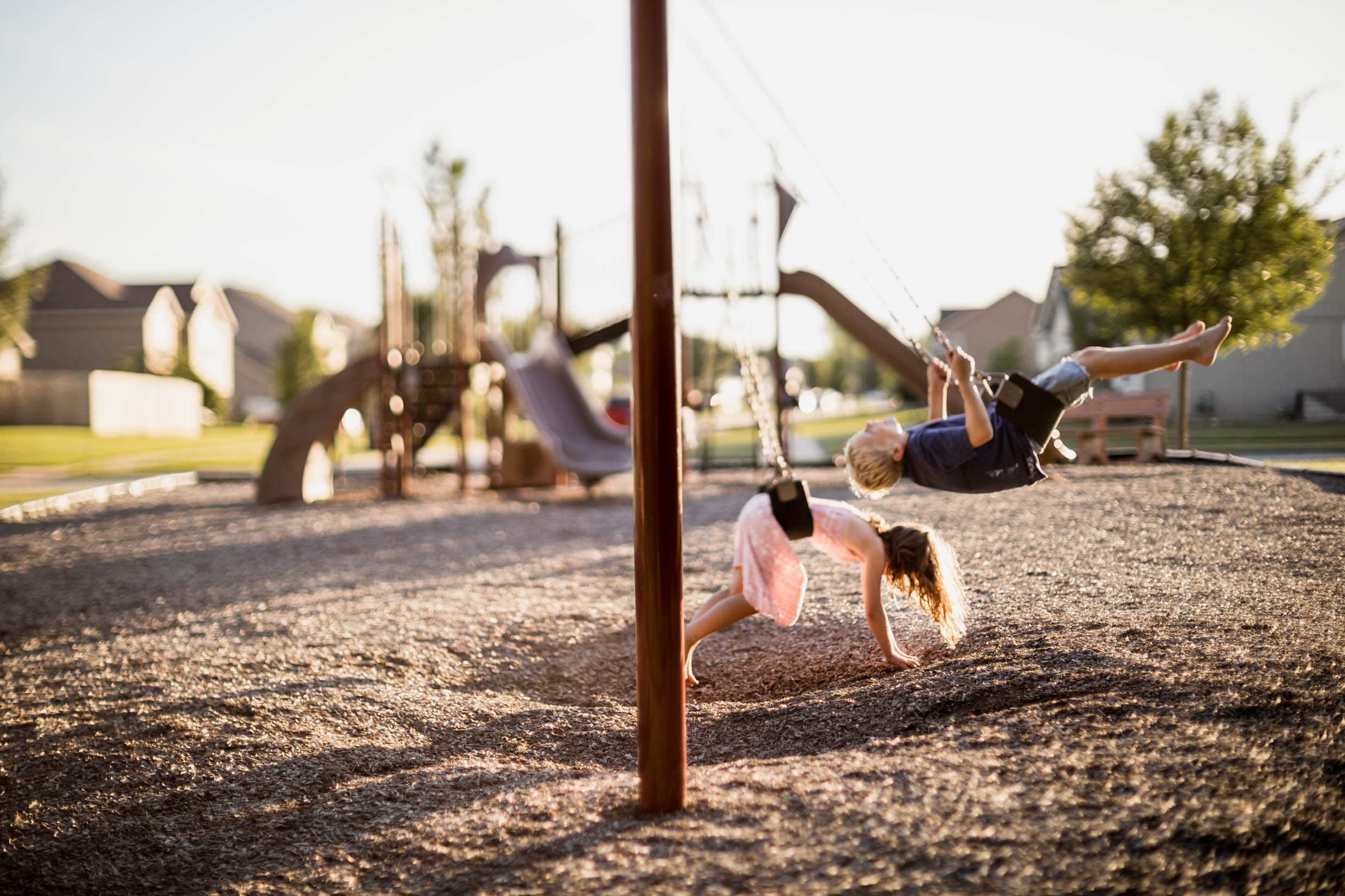Child swinging on a playground with a person falling nearby, in a neighborhood setting with houses and trees, during daytime.