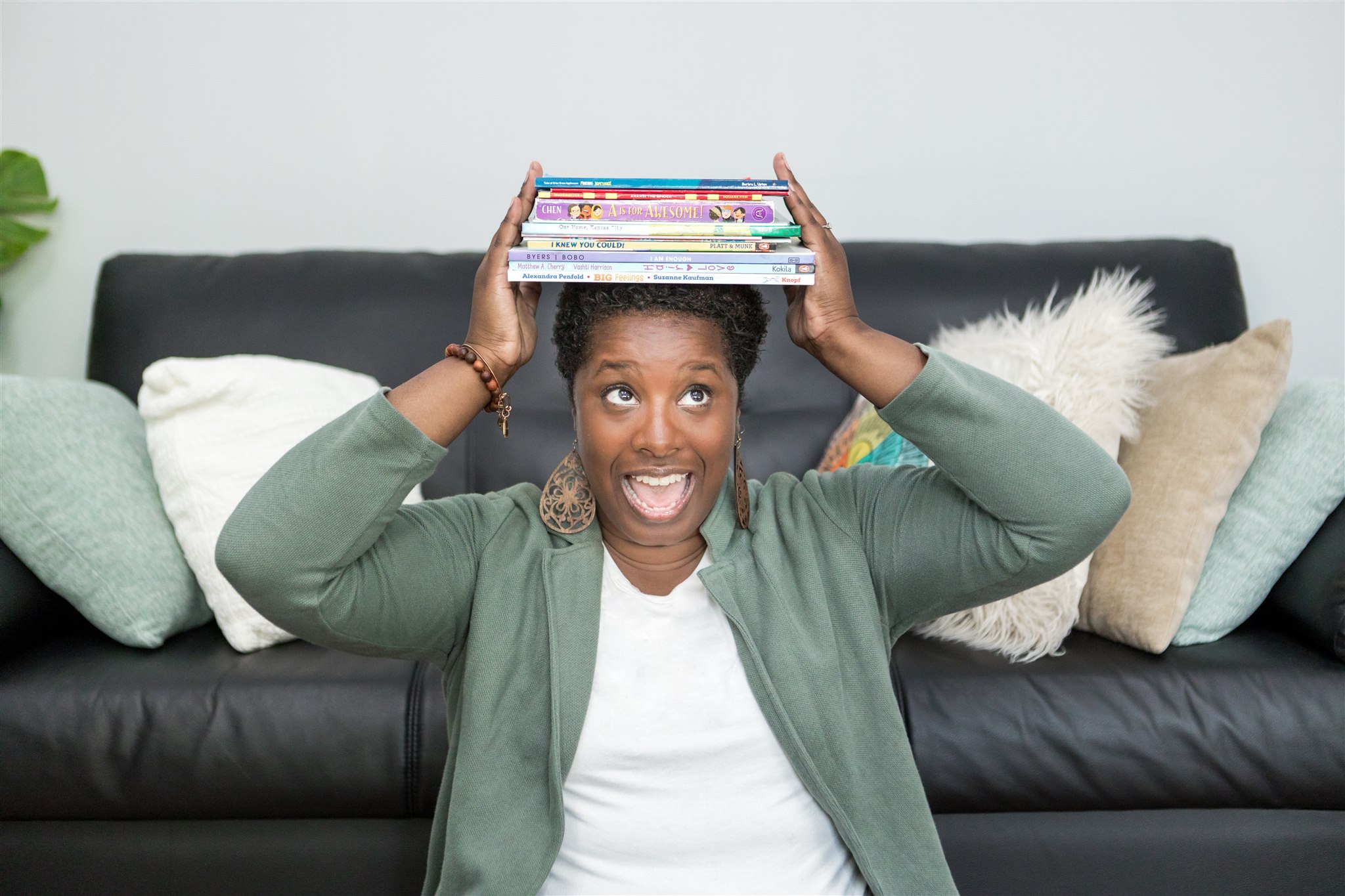 Woman with short hair and a green jacket balances a stack of books on her head, smiling in a living room.