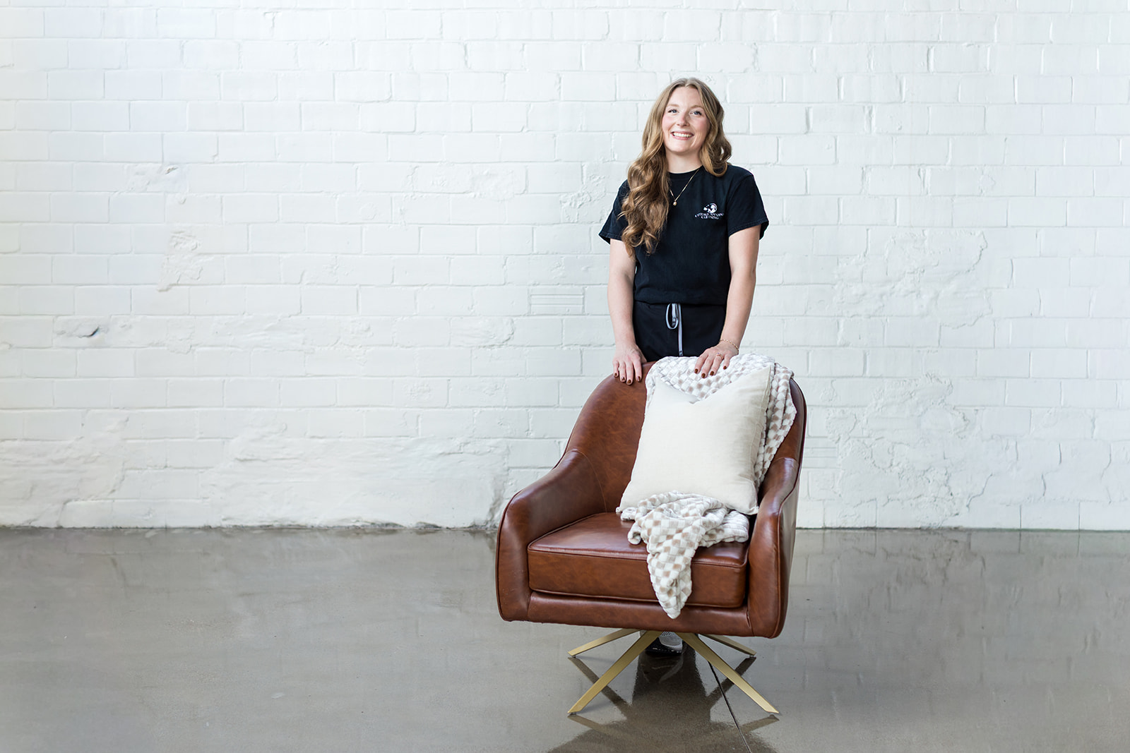Young girl with long hair standing behind a brown armchair with a blanket draped over it, against a white brick wall.