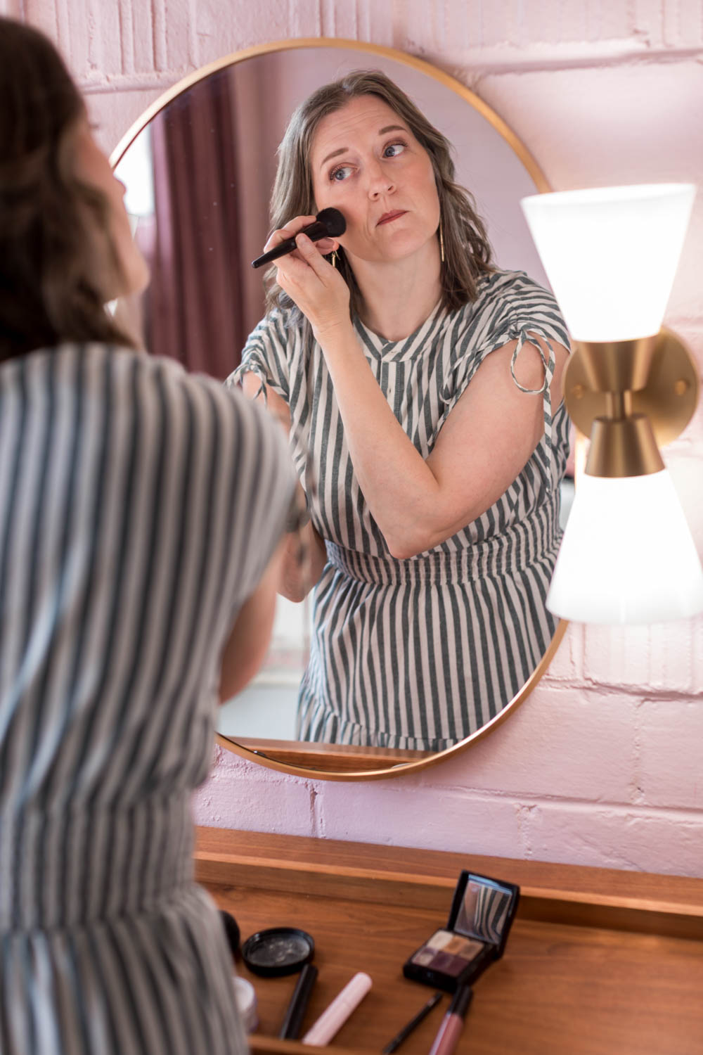 Woman applying makeup while looking in a mirror, with a lamp on the side, on a pink wall background.