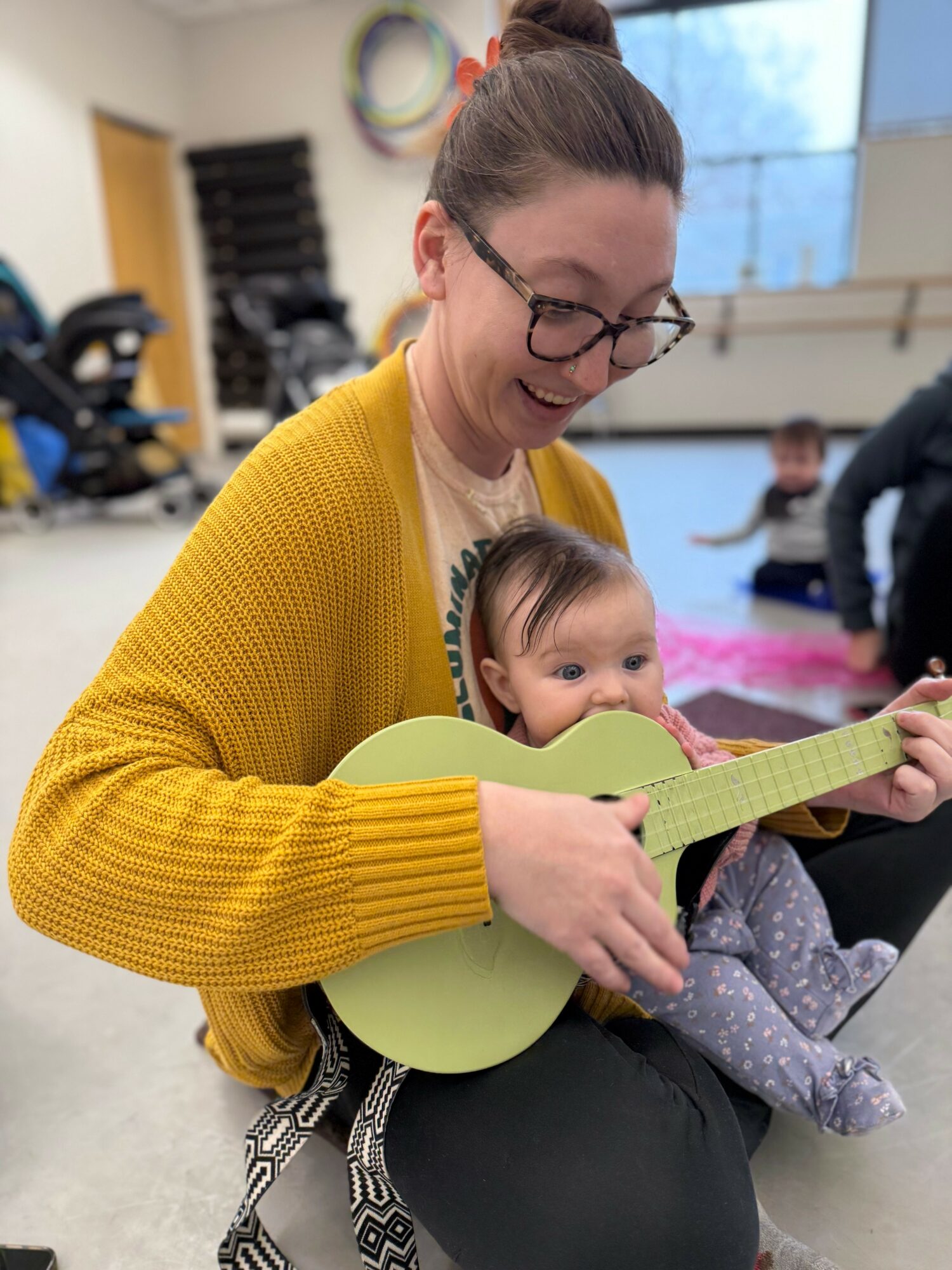 Woman with glasses and a yellow sweater holds a child with a green guitar, smiling indoors.