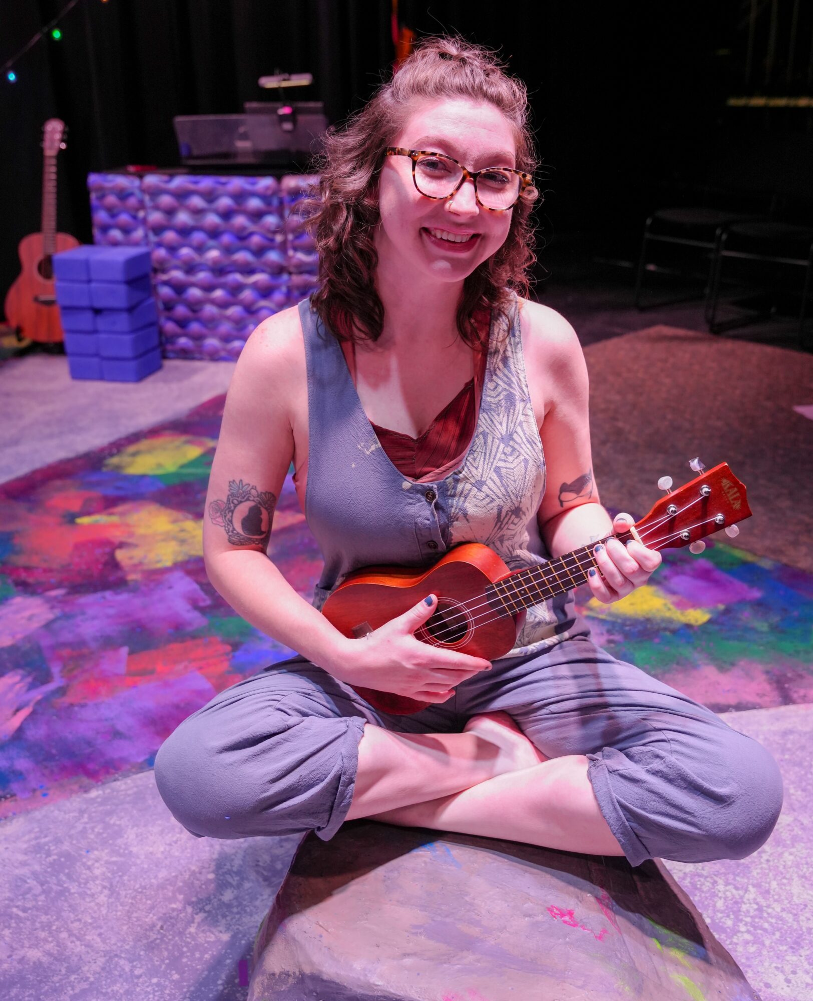 Person sitting cross-legged on colorful stage holding a ukulele, smiling, with musical equipment in background.