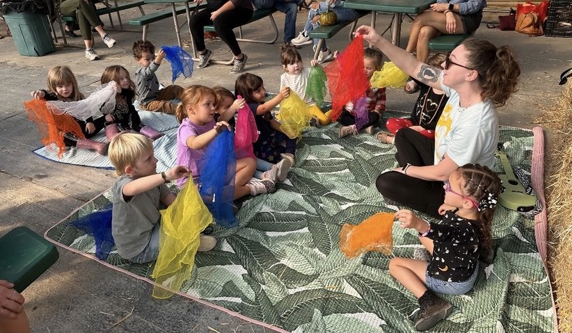 Children sitting on a patterned mat, holding colorful scarves, with an adult woman leading activity outdoors.