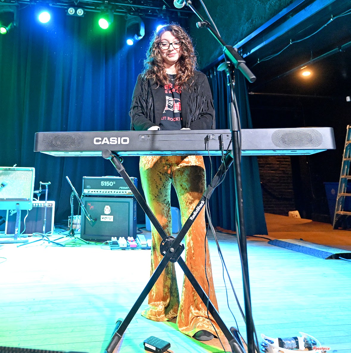 Woman with curly hair and glasses standing behind a keyboard on stage, smiling, with stage lights and equipment in background.