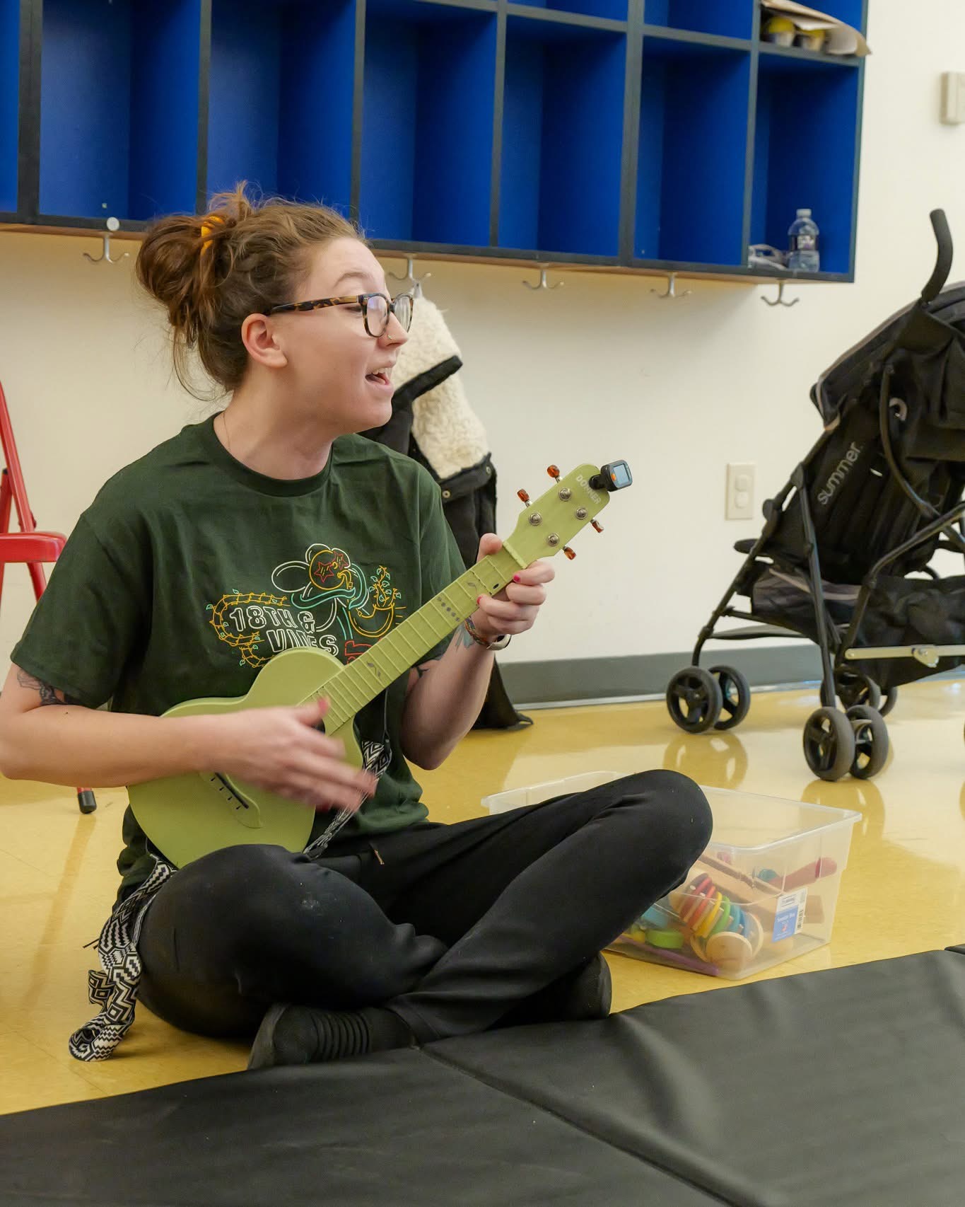 Person sitting cross-legged on the floor playing a small yellow guitar, with a stroller and blue storage cubbies in the background.