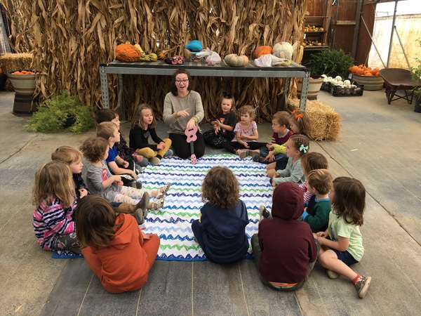 Group of children and an adult sitting on a blanket in a barn-like setting with hay bales and pumpkins, participating in a group activity.