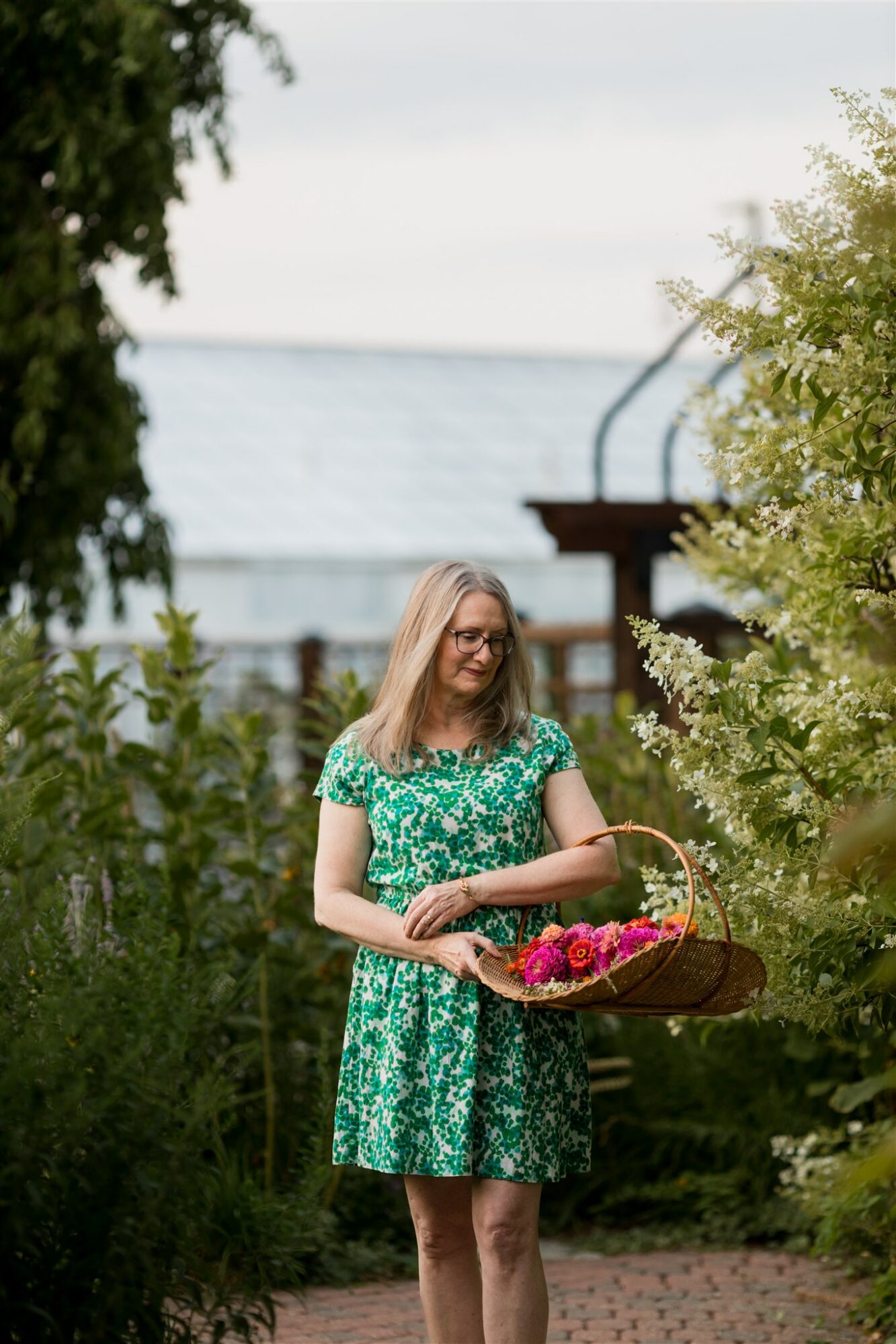 Woman in green floral dress holding a basket of flowers outdoors, surrounded by trees and plants.