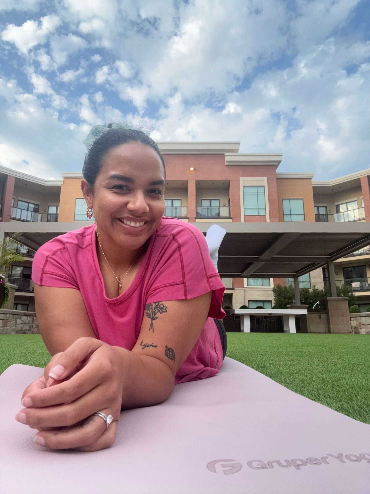 Woman in pink shirt smiling, leaning on table outdoors with modern buildings and sky in background.