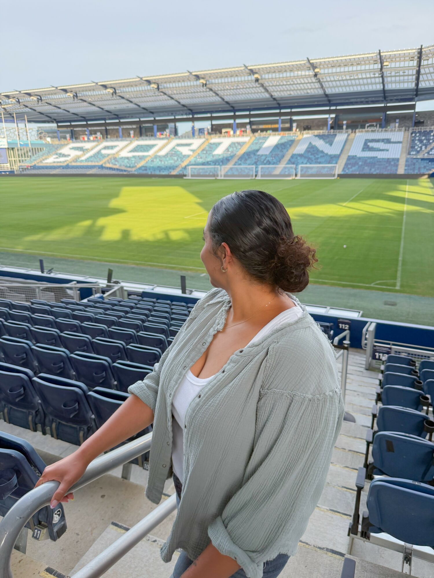 Woman with dark hair tied back, wearing a light gray jacket, sitting in stadium seats overlooking a sports field.