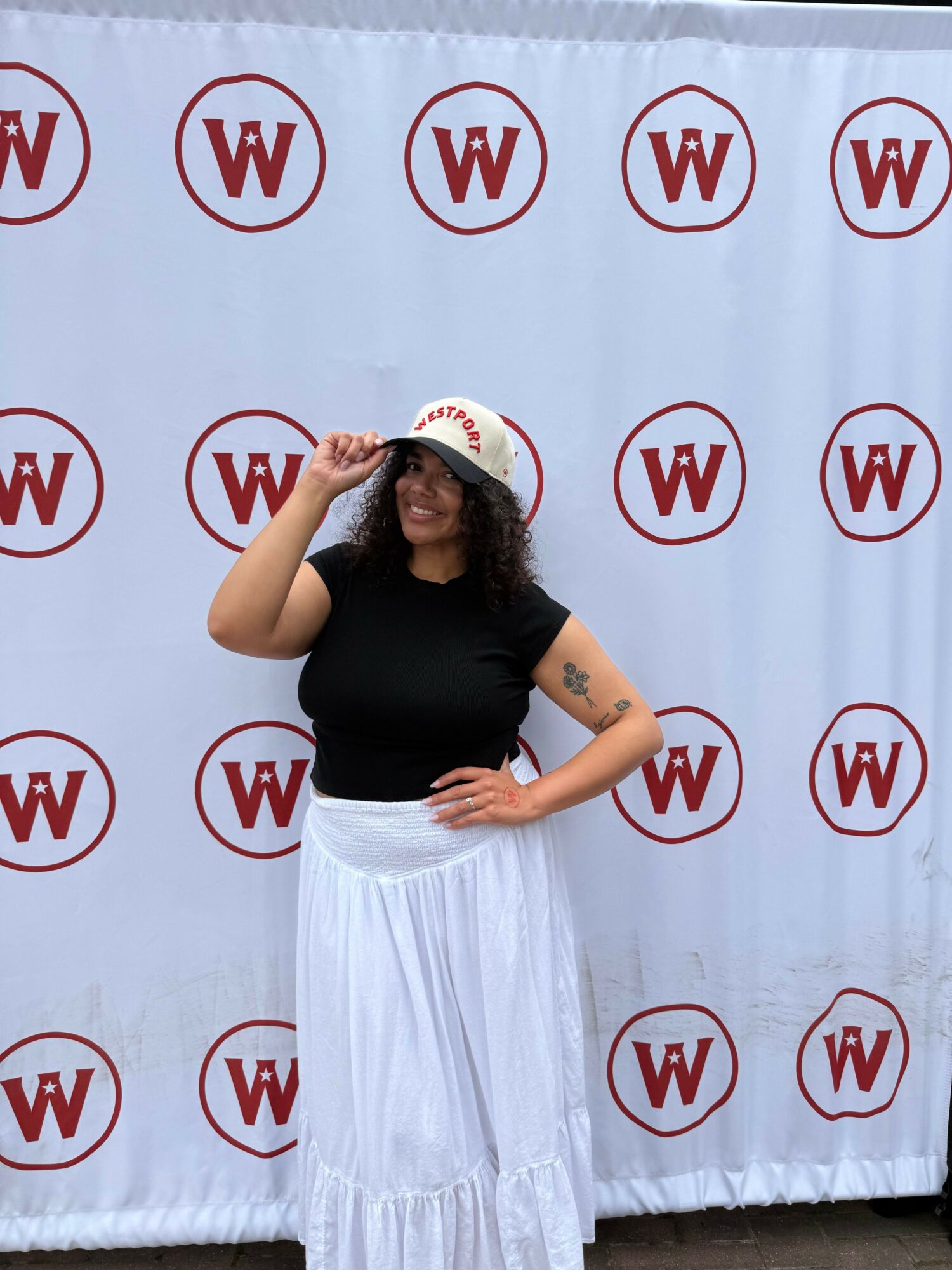 Person with long hair wearing a black shirt and white skirt, standing in front of a backdrop with red 'W' logos.