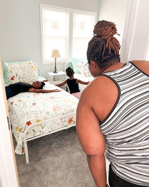 Woman with braided hair and striped tank top looking at two children in a bedroom with a bed, window, and lamp.