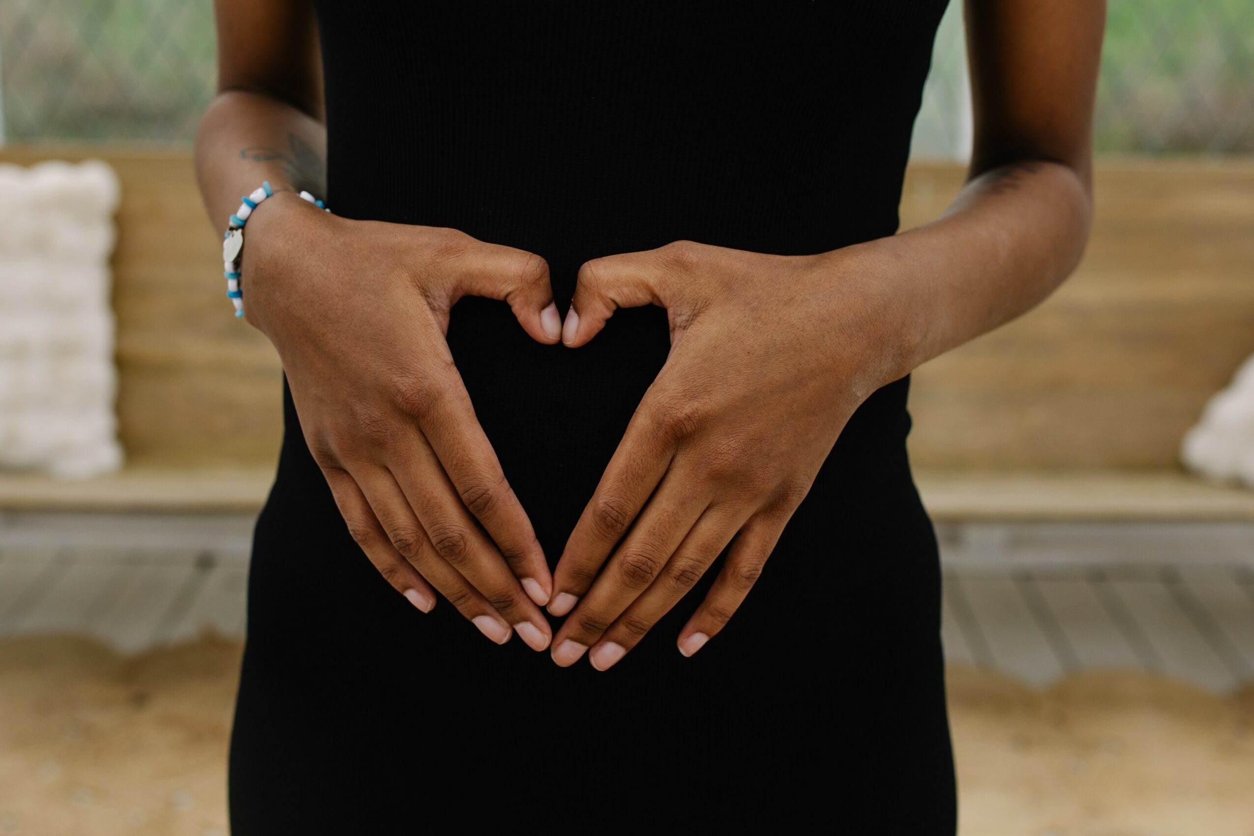 Person forming a heart shape with hands on stomach, wearing a black dress, in a room with wooden background.