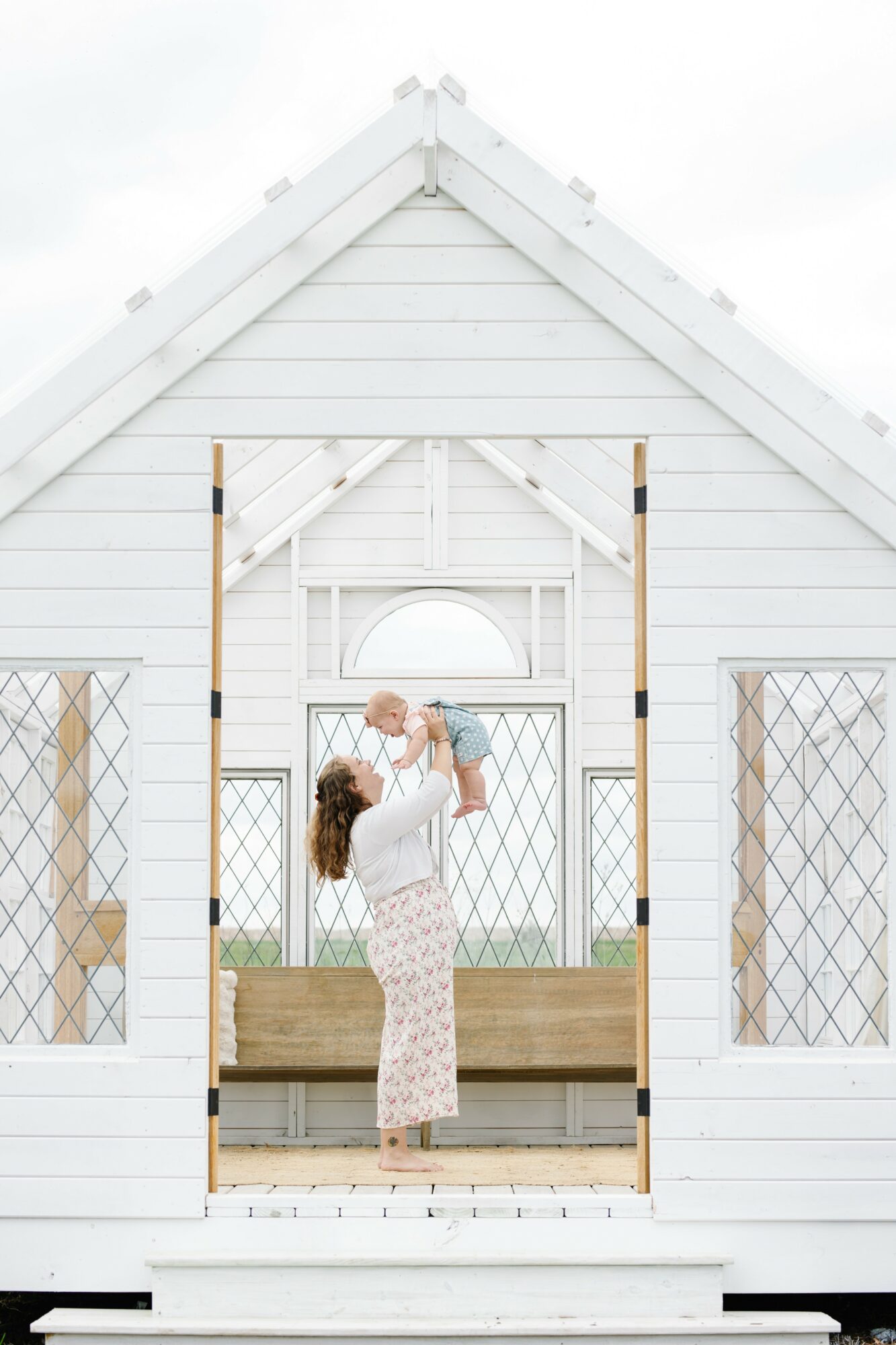 Woman standing on porch, holding a child, in front of a white house with large windows and a triangular roof.