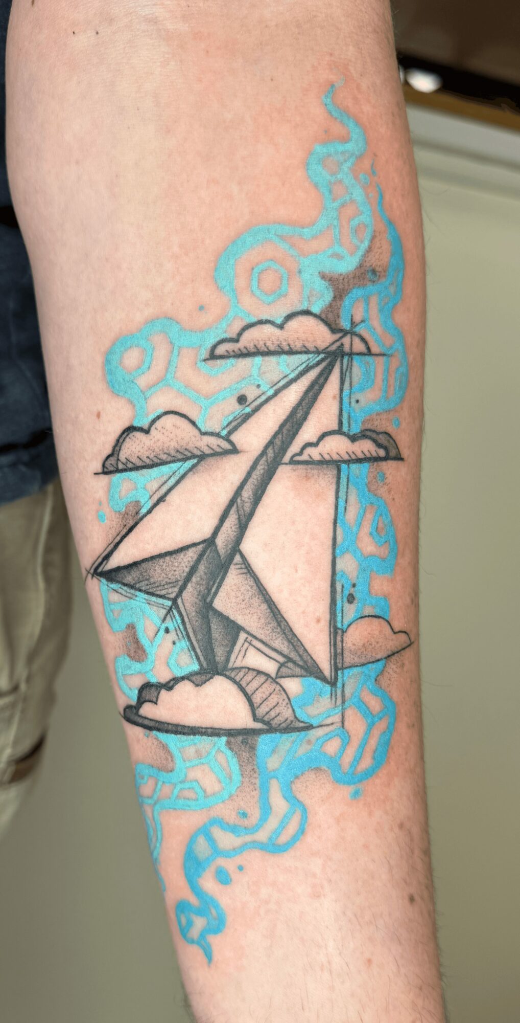 Tattoo of a paper airplane flying through clouds with blue swirling background on person's arm.