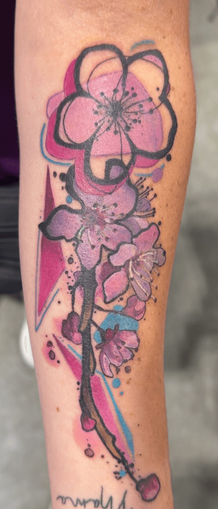 Colorful flower tattoo with pink, purple, and blue hues on a person's arm, with abstract splashes and lines.