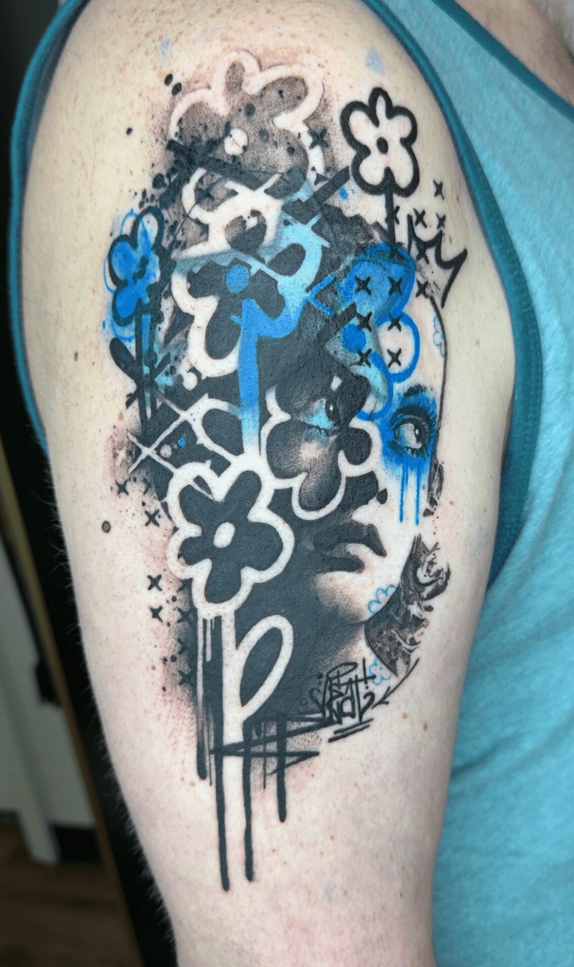 Tattoo on upper arm with flowers, abstract shapes, and a face, in black, blue, and pink colors.