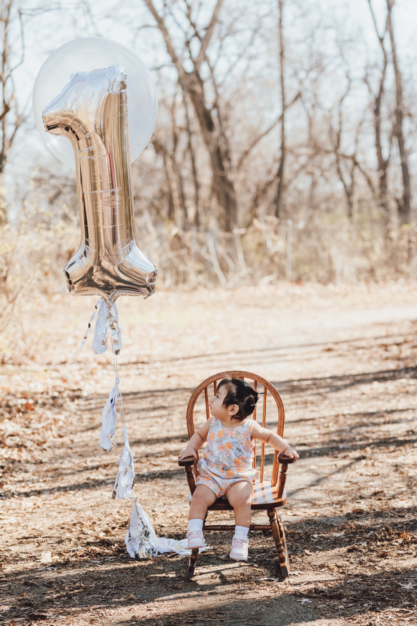 A young girl sitting on a wooden chair outdoors with a large silver number 1 balloon nearby, trees in the background.