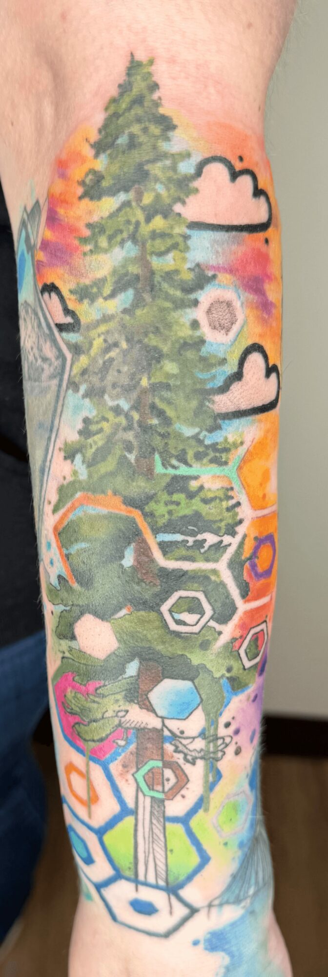 Colorful tattoo of trees, clouds, and geometric shapes on a person's arm, with vibrant sunset colors in the background.