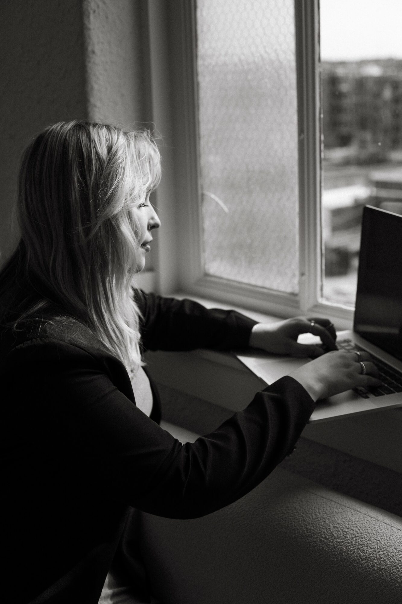 Woman working on a laptop by a window, looking outside, in black and white.