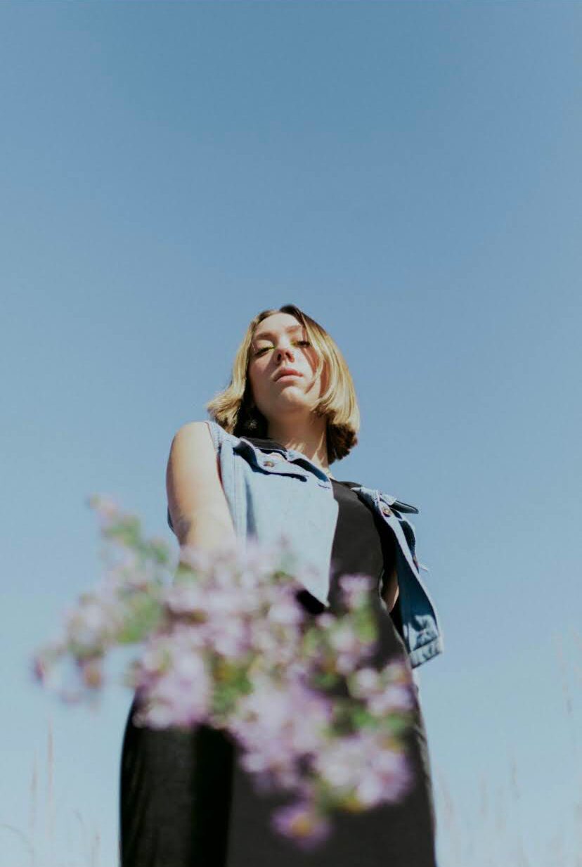 Young woman with shoulder-length hair stands outdoors against a clear blue sky, with blurred pink flowers in foreground.