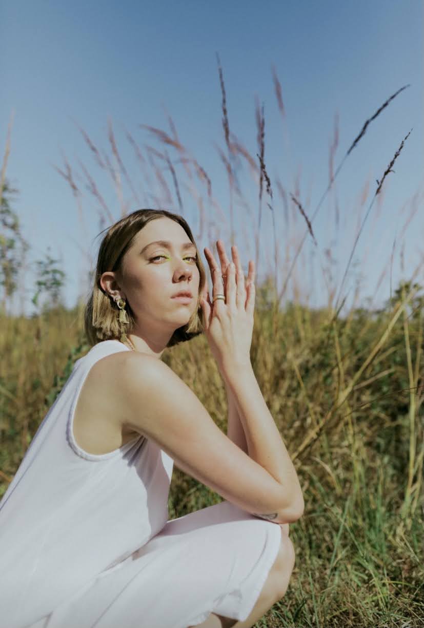 Young woman with short hair and earrings in a white dress outdoors, with tall grass and a blue sky behind her.