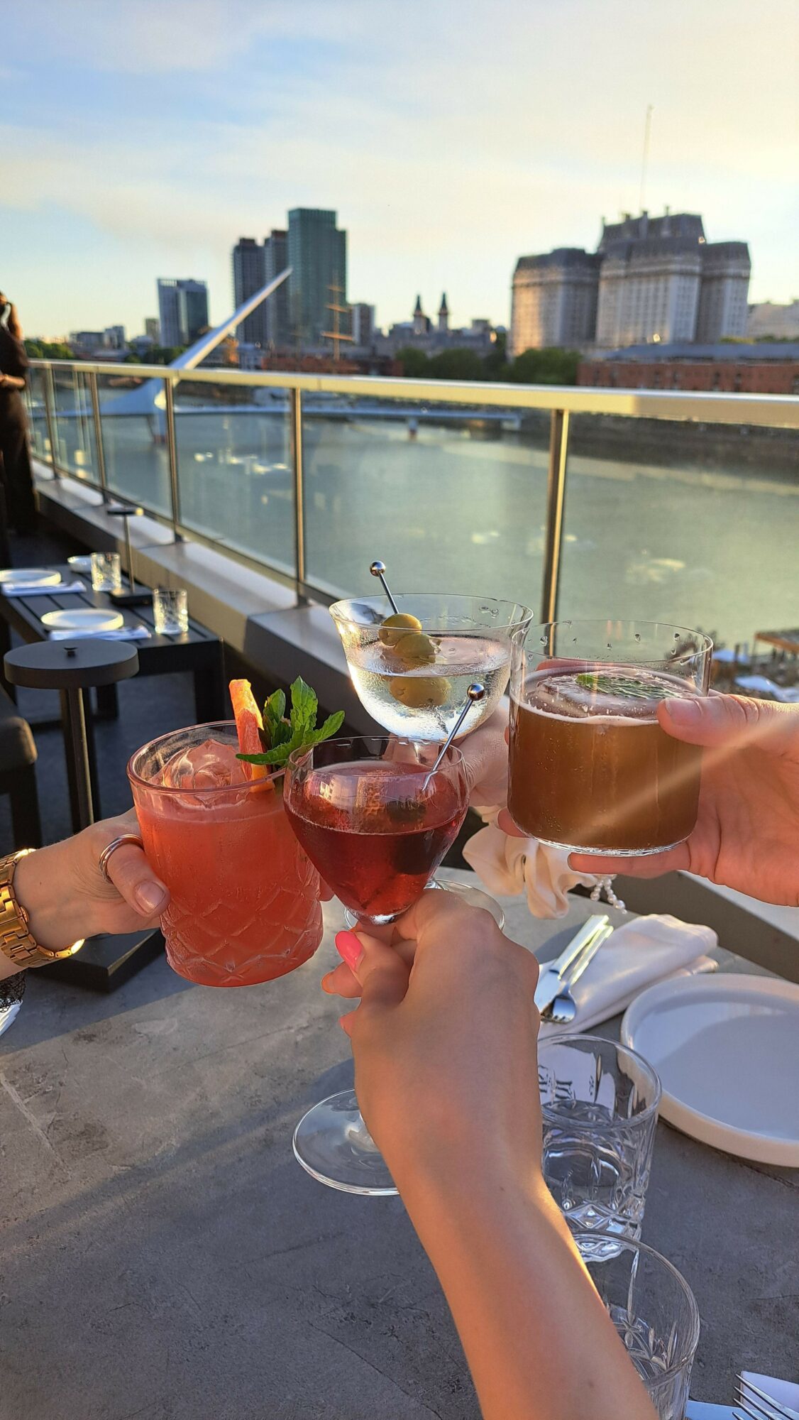 People toasting with drinks on a rooftop overlooking a city skyline and river, during sunset.