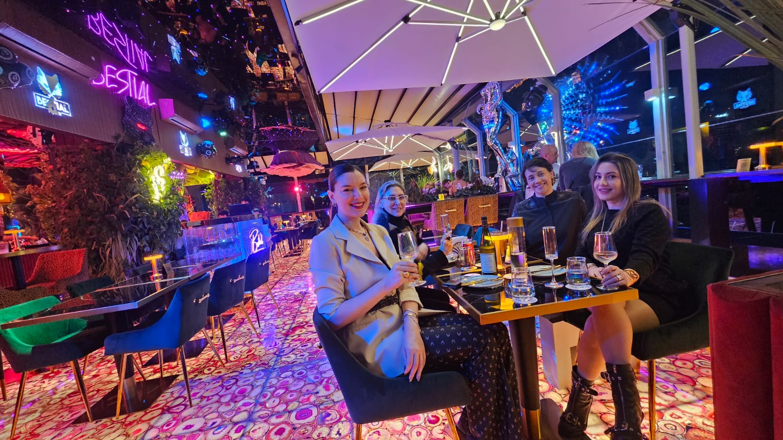 Three women sit at a table in a colorful, neon-lit outdoor restaurant or bar at night, with umbrellas overhead.