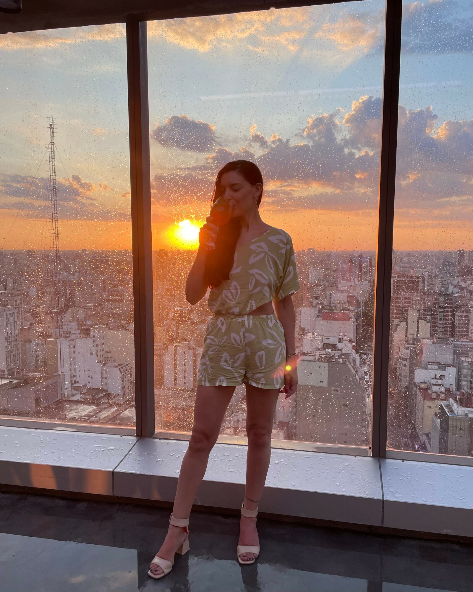 Person standing inside a high-rise building with large windows, cityscape and sunset in background, wearing a patterned outfit and high heels.