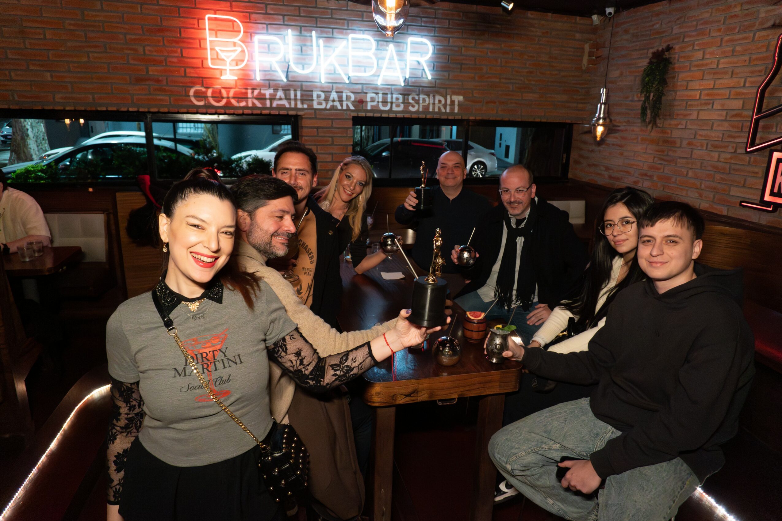 Group of people sitting around a bar table in a pub, smiling and holding drinks, with neon signs in the background.