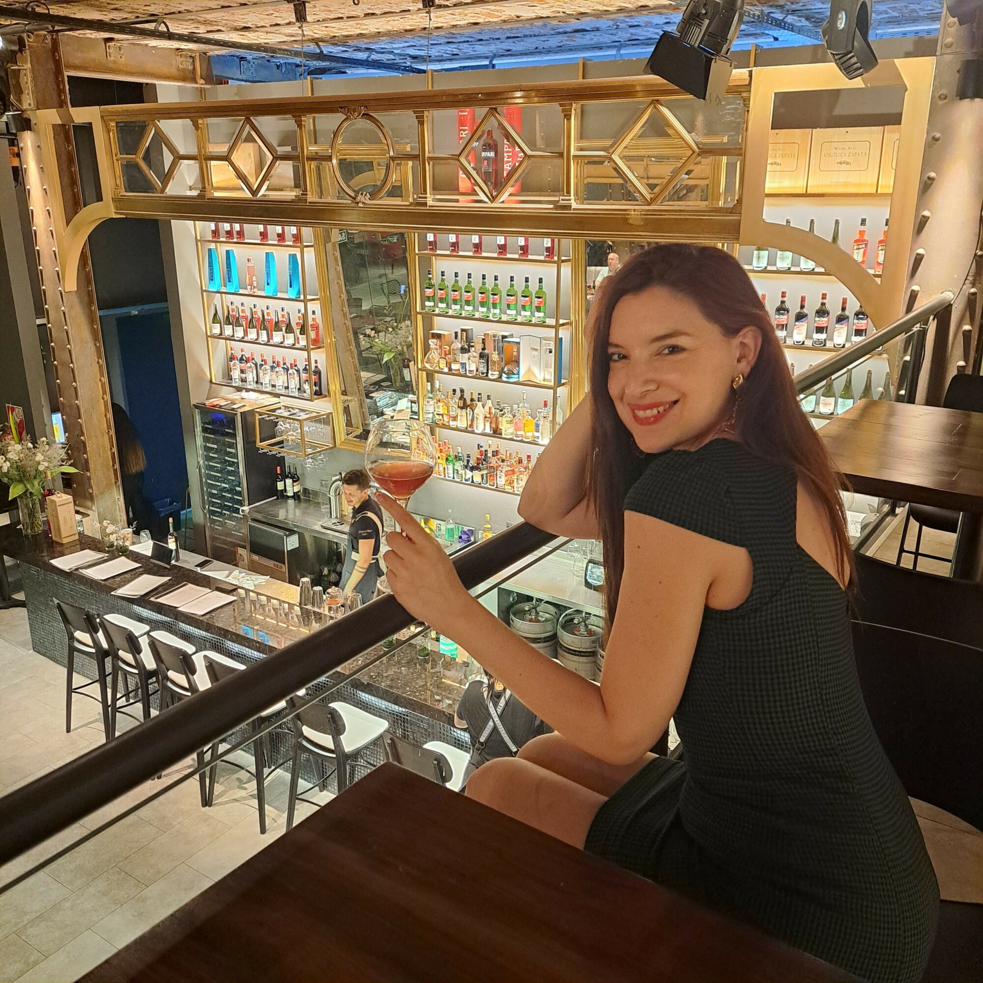 Woman with long hair leaning on a bar counter, smiling, in a brightly lit bar or restaurant setting.