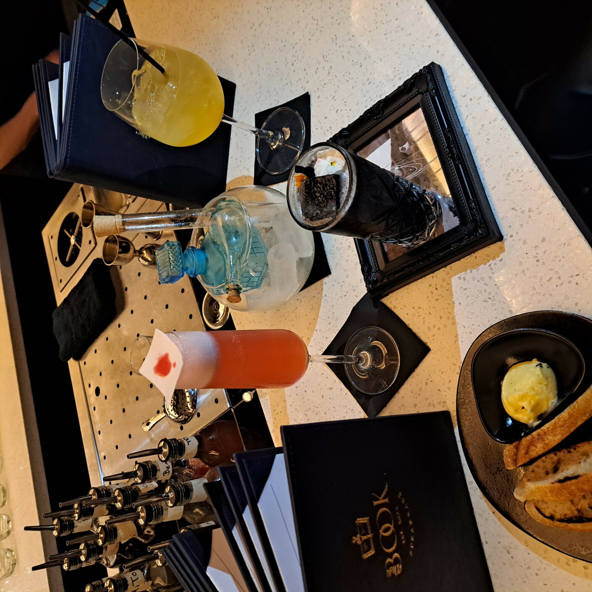 Table with various drinks, a framed photo, and snacks, viewed from a tilted angle. A black menu is visible.