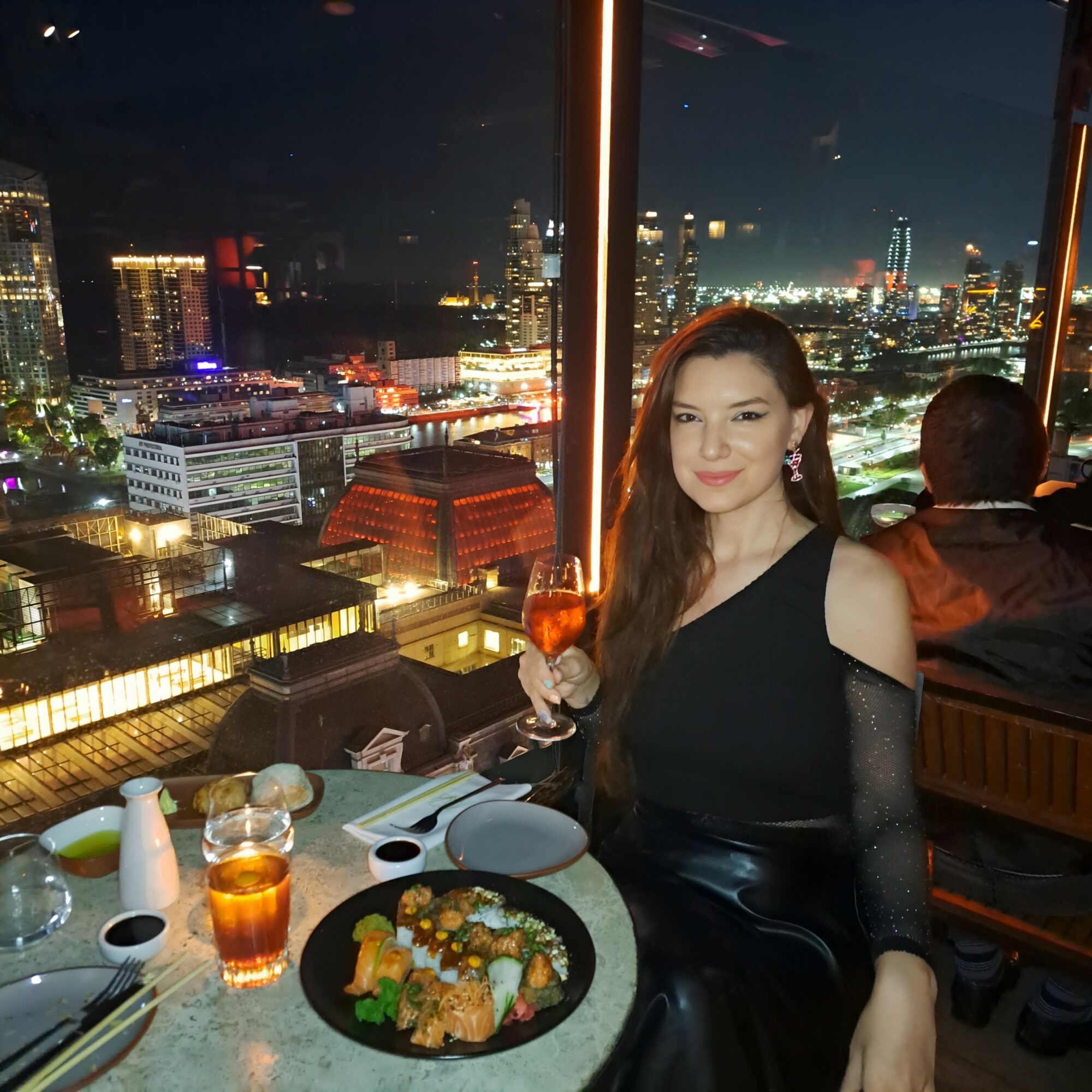 Woman in black dress holding a glass at a high-rise restaurant with city skyline at night, sushi on table, city lights visible.