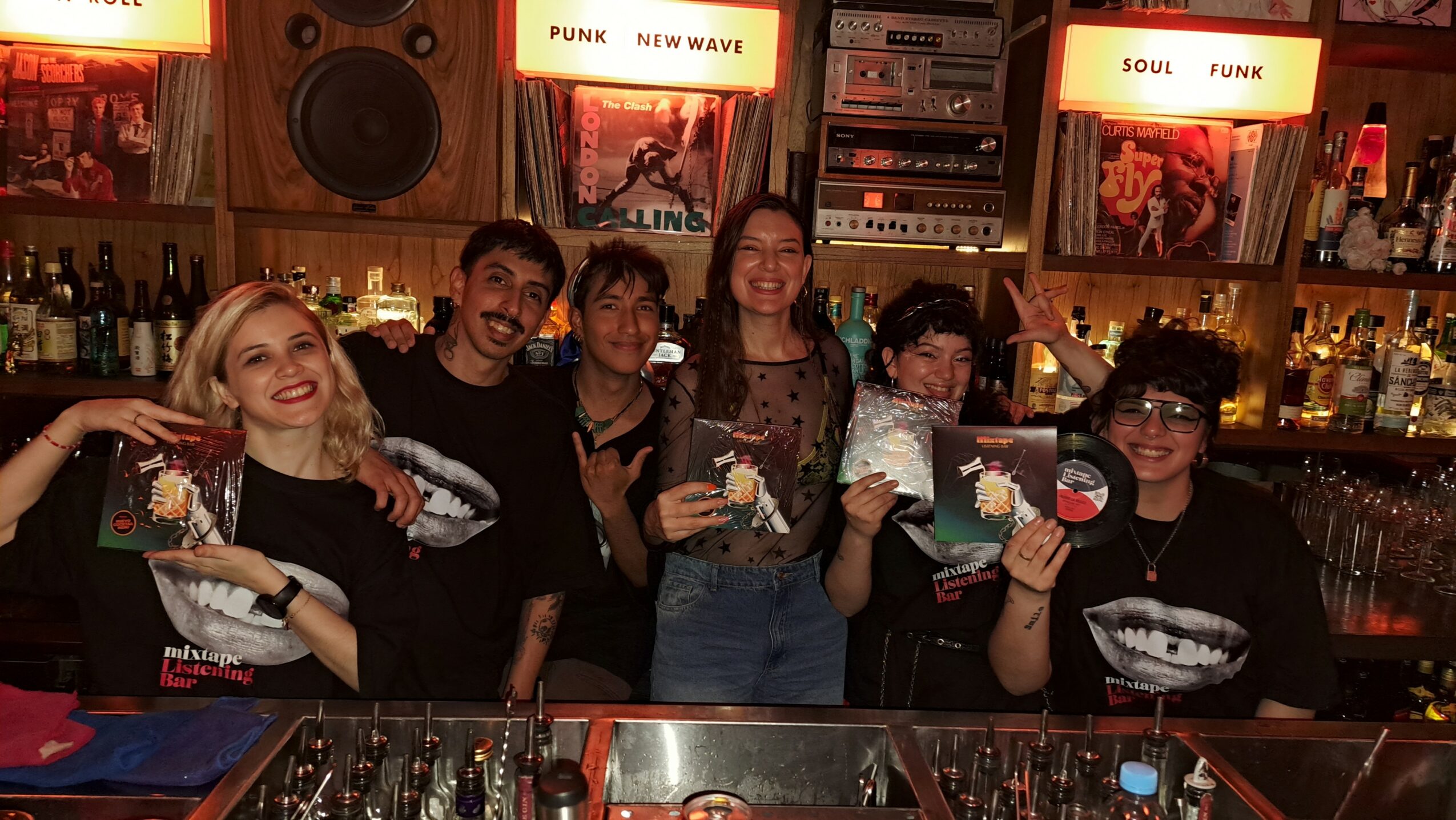 Group of six people behind a bar, holding food and drinks, smiling, with bar equipment and illuminated signs in the background.