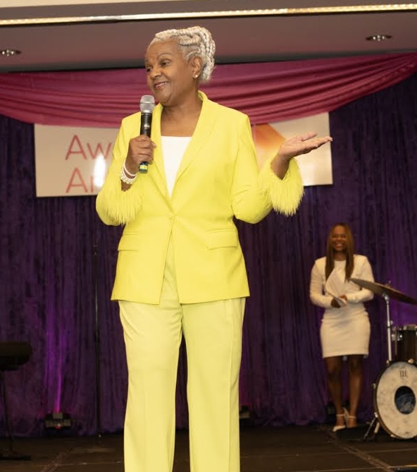 Elderly woman with short gray hair speaking into a microphone on stage, wearing a bright yellow suit, with a woman in the background. Word partially visible on banner.