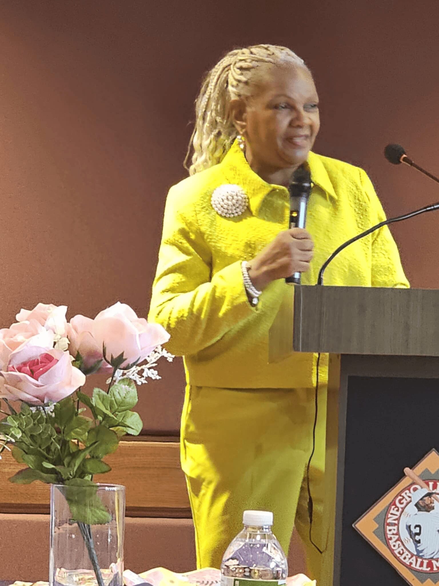Woman in yellow suit speaking at a podium with a microphone, pink roses in a vase nearby, and a water bottle.