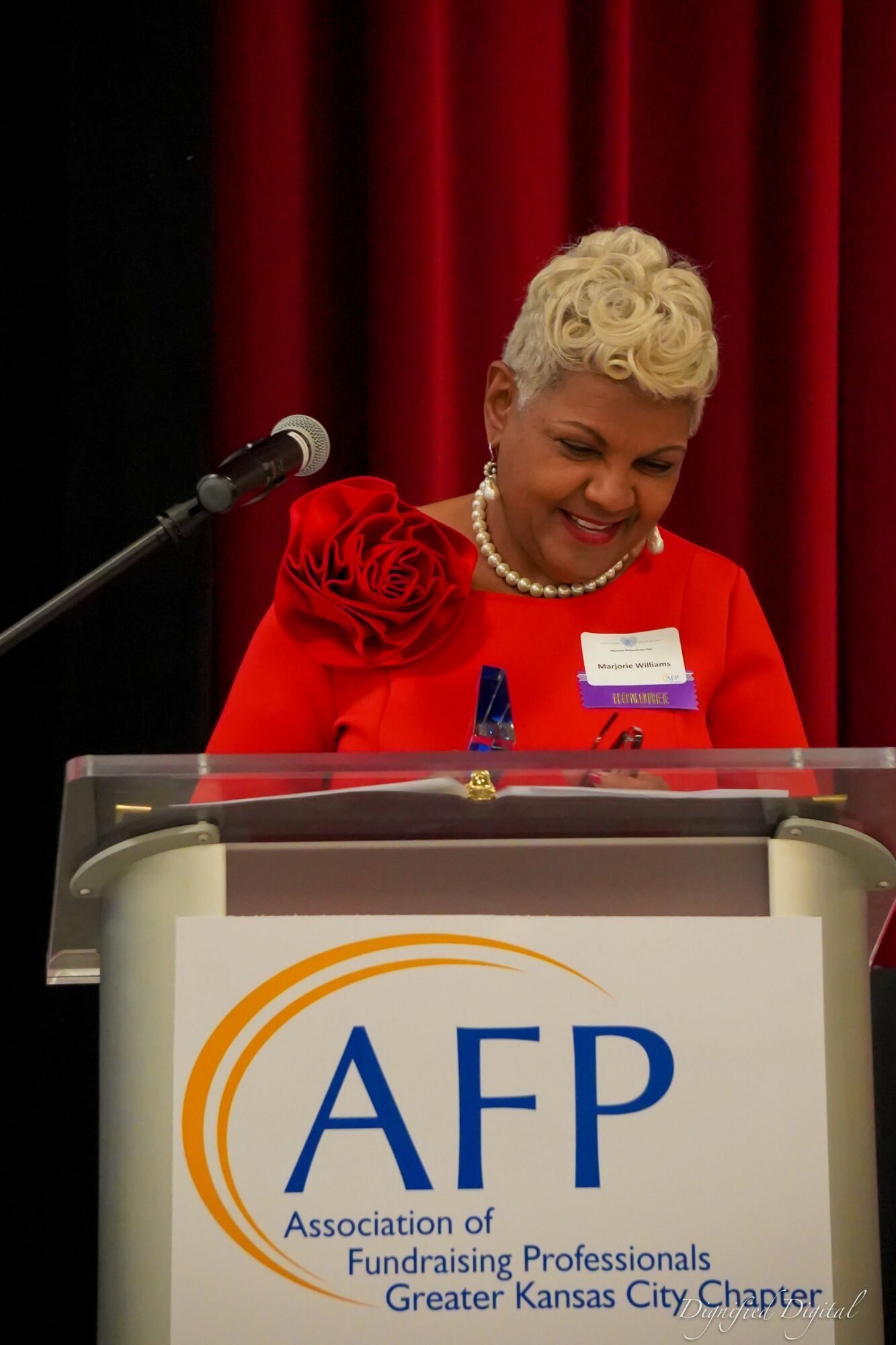 Woman with short, curly blonde hair wearing a red dress with floral details, speaking at a podium with a red curtain background.