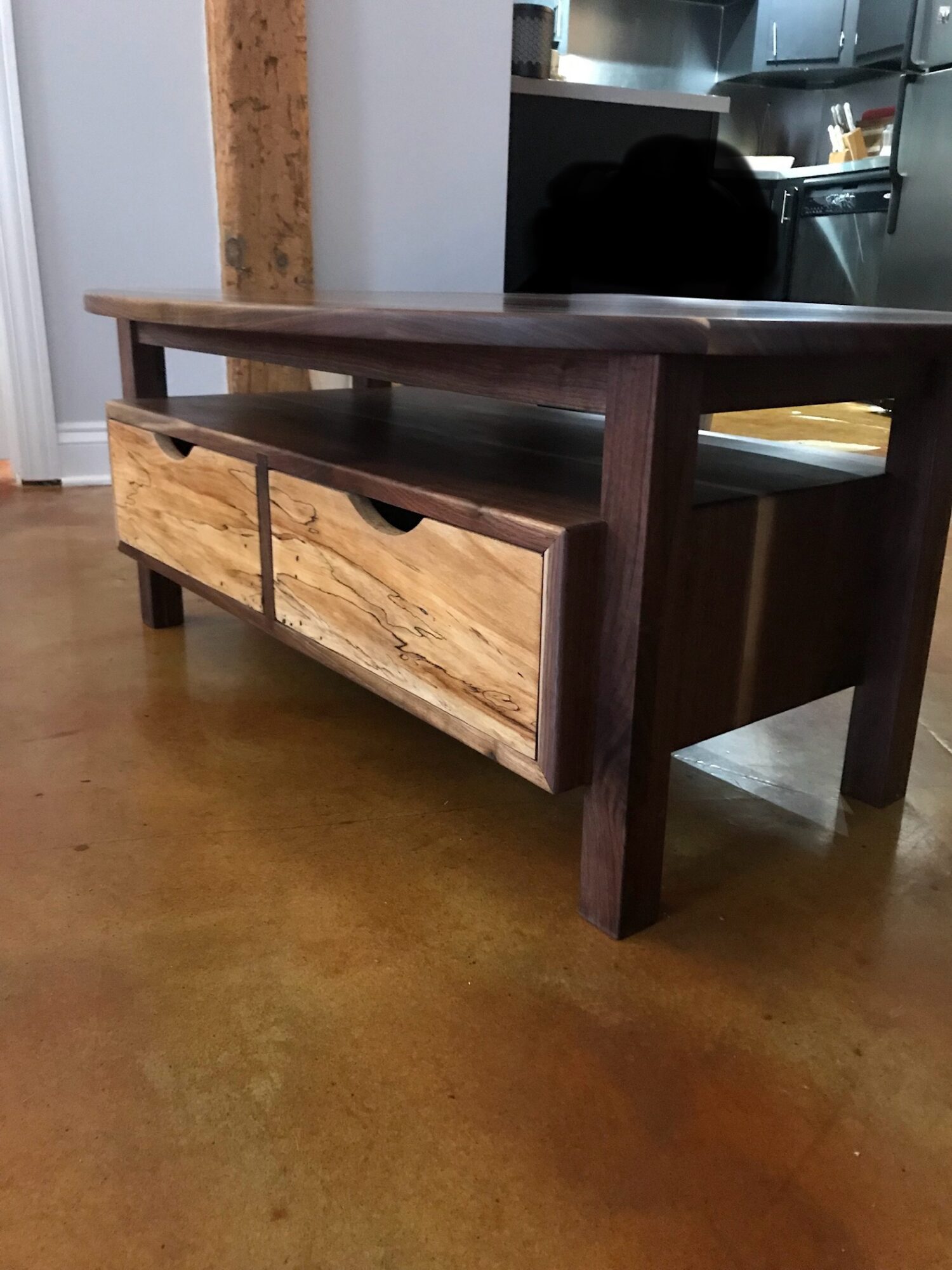 Wooden table with two drawers, placed on a wooden floor, with a kitchen in the background.