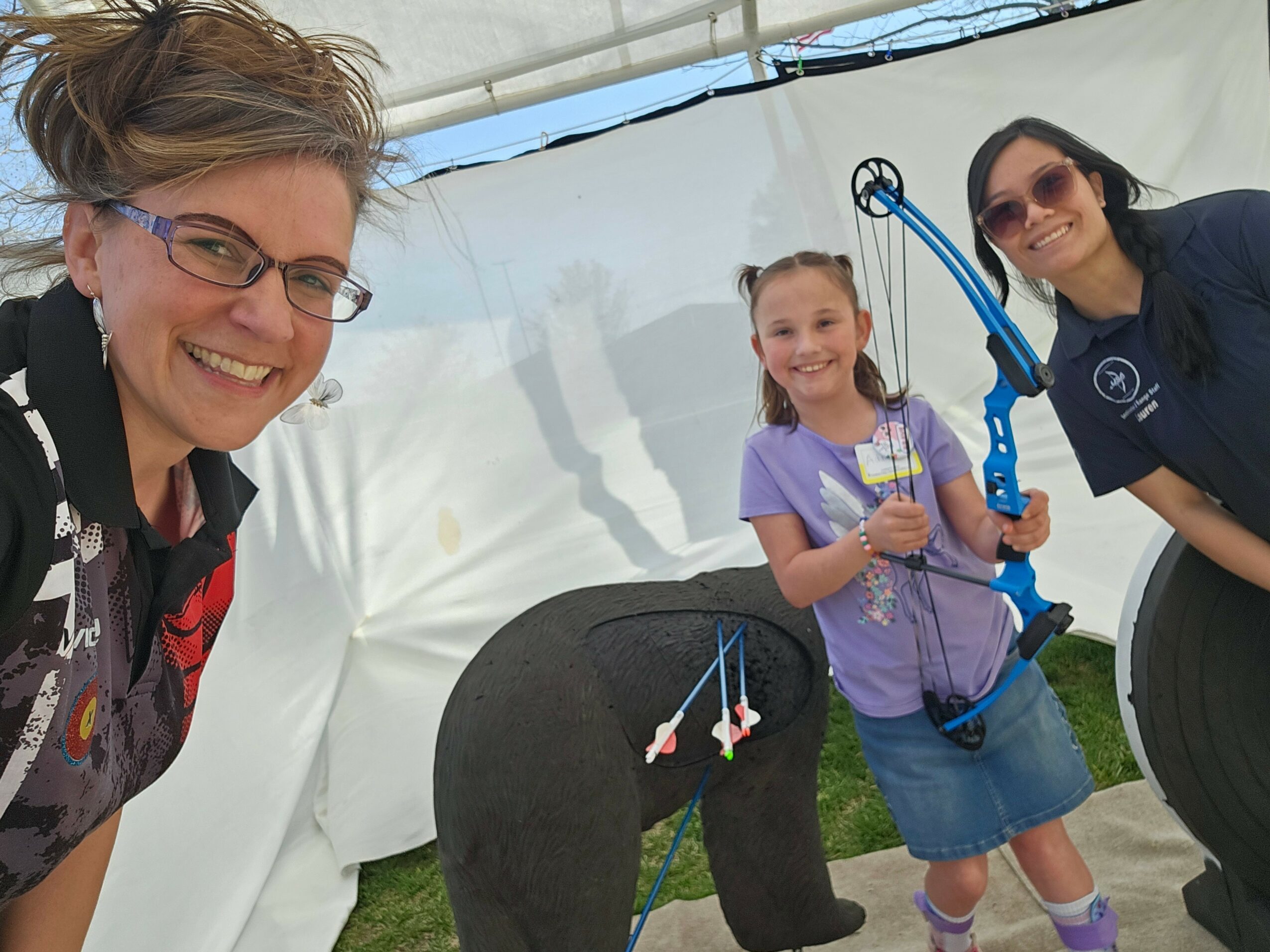 Three smiling people, two women and a girl, inside a tent, holding archery equipment and a trophy.