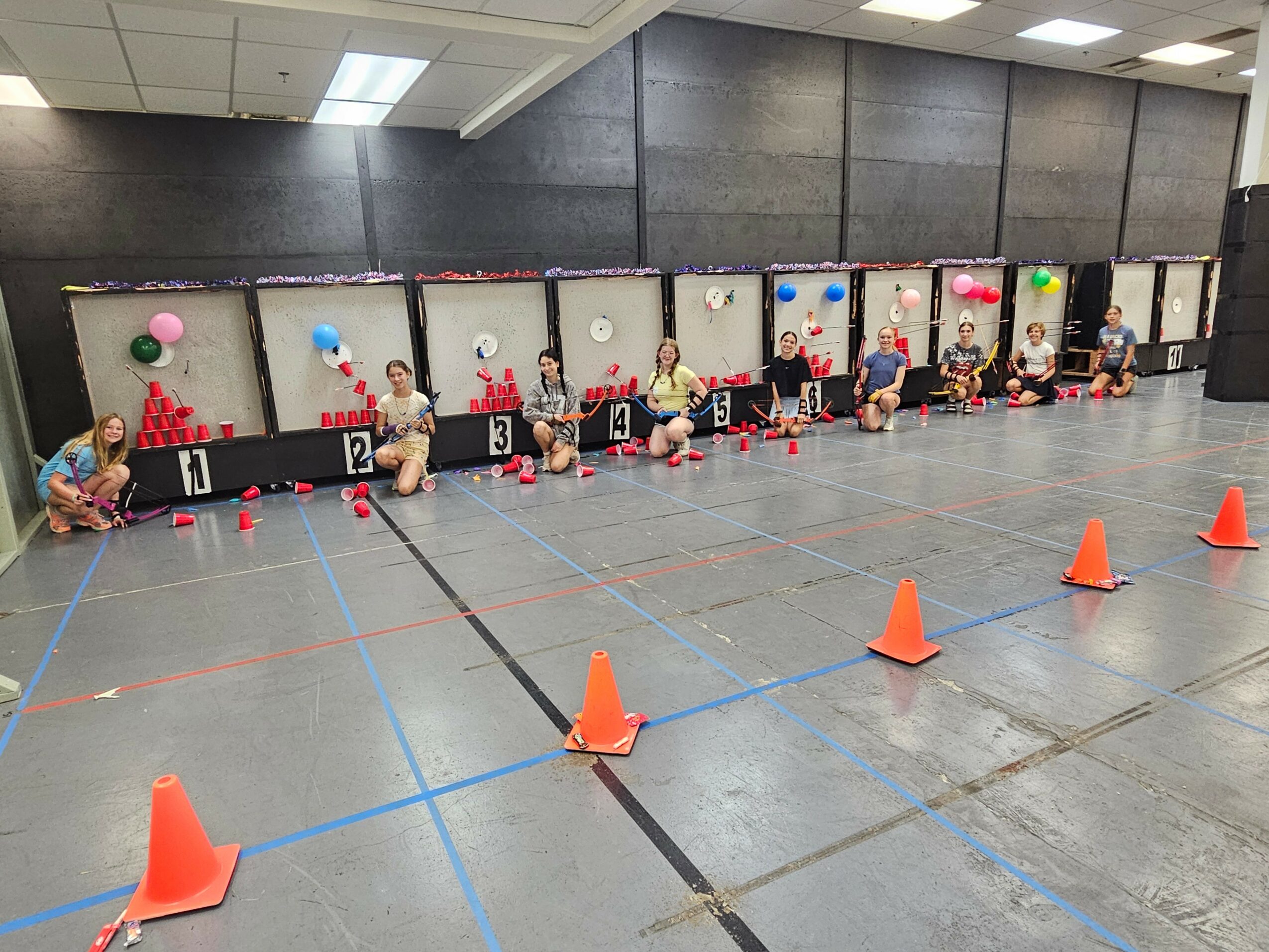 Children sitting on the floor in front of cubicles decorated with balloons, with orange cones in the foreground, in an indoor space.