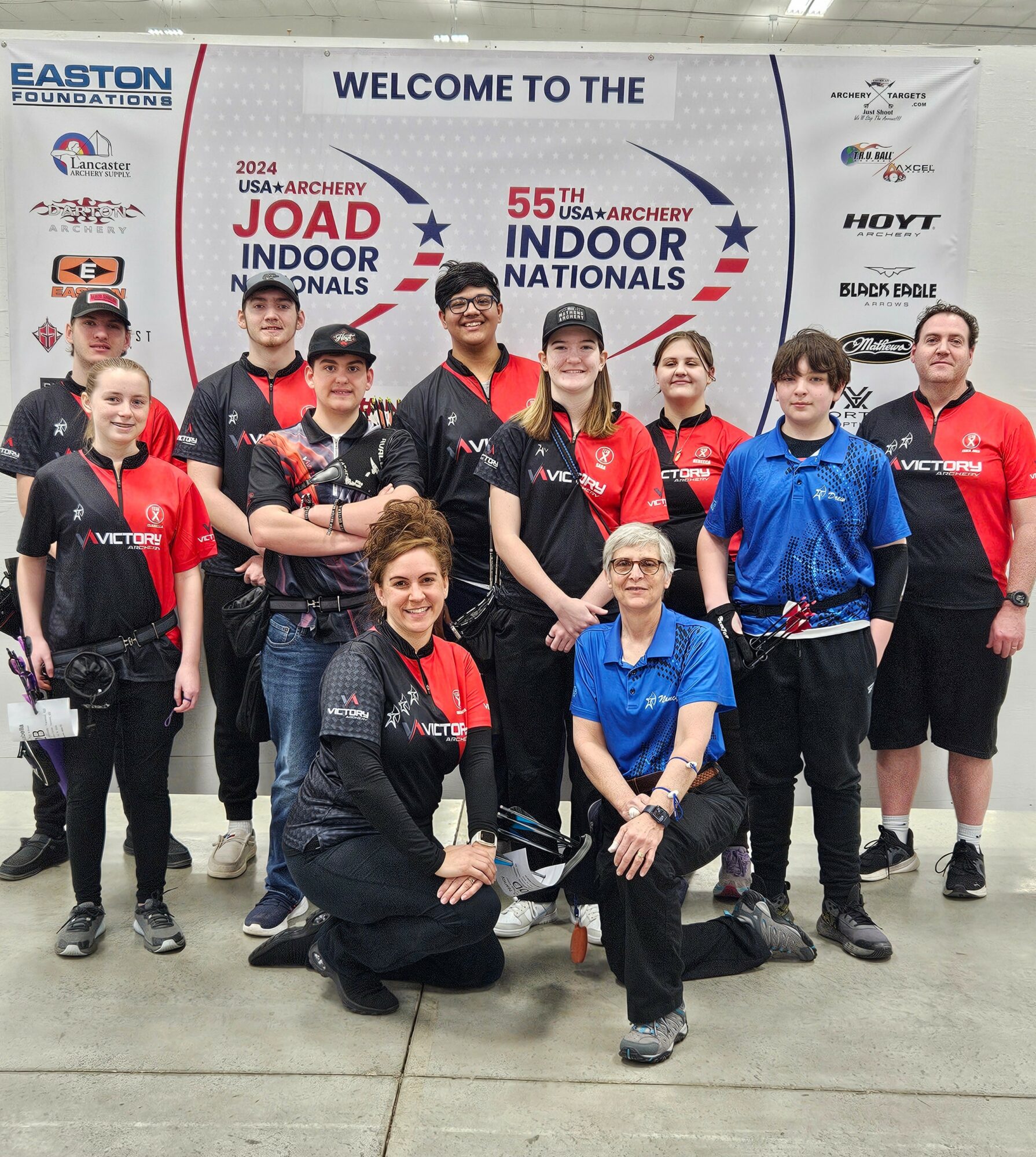 Group of ten people, including children and adults, standing in front of a banner at an indoor archery event, smiling.