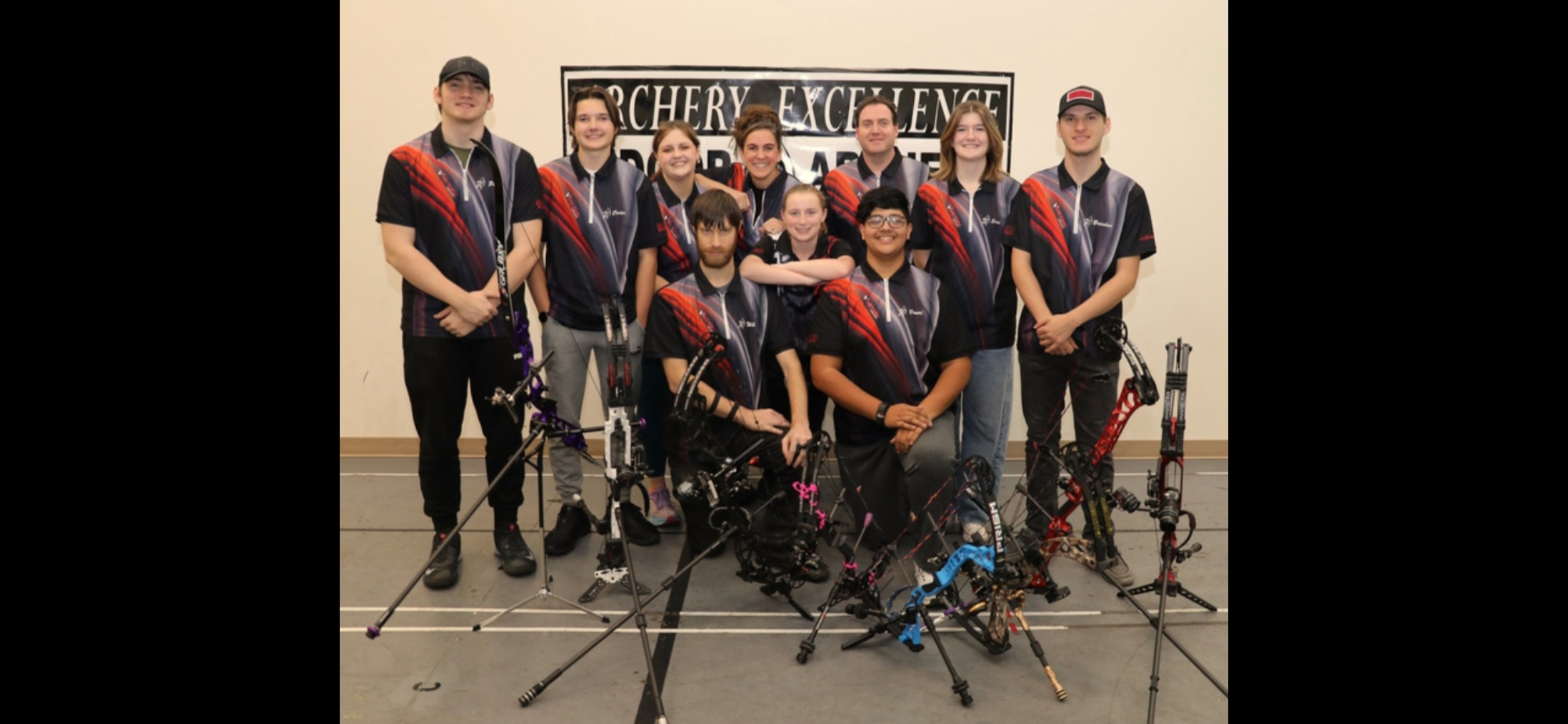 Group of archers in uniform posing with bows and arrows indoors, with a sign in the background.