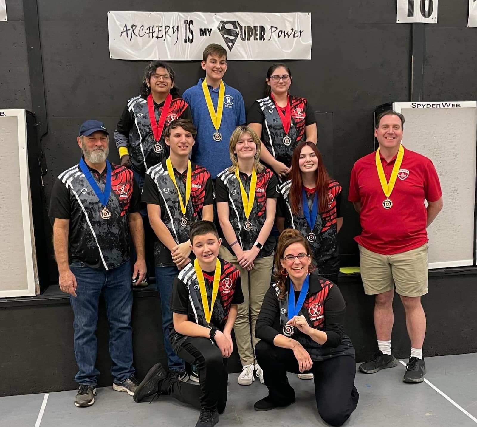 Group of nine people, some wearing medals, standing indoors in front of a black wall with a banner. They are smiling.