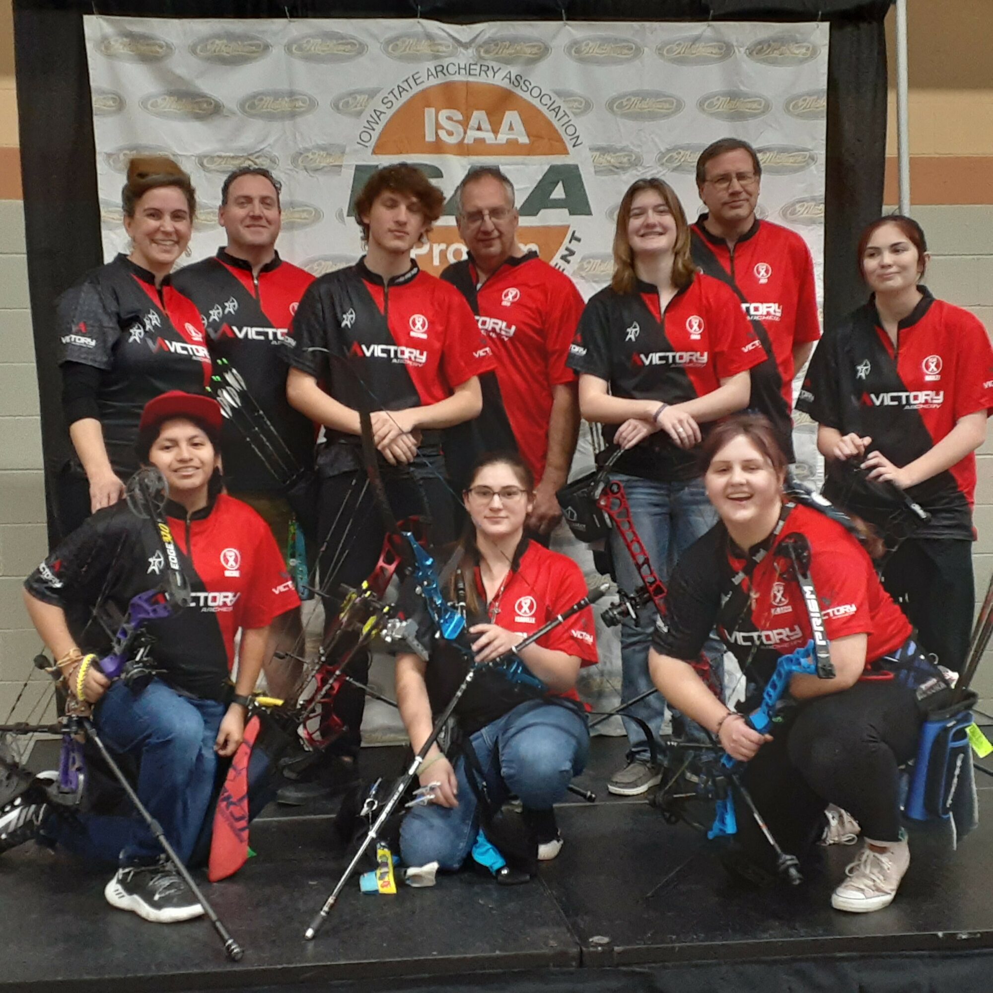 Group of ten people posing with archery equipment in front of a banner, some wearing black and red shirts, some with glasses, smiling.