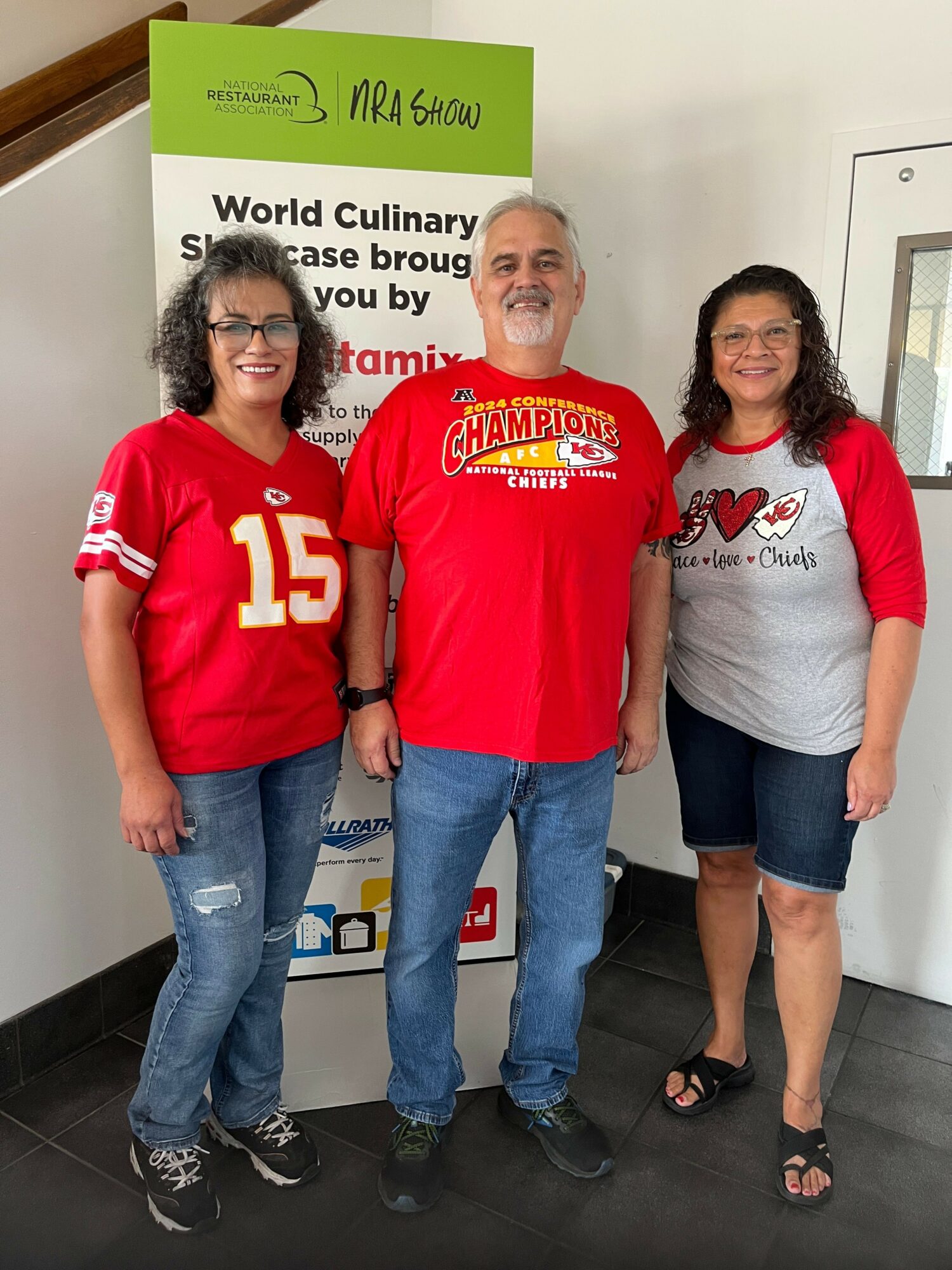 Three women standing indoors near a sign, one wearing a red sports jersey, two in casual clothing, smiling.