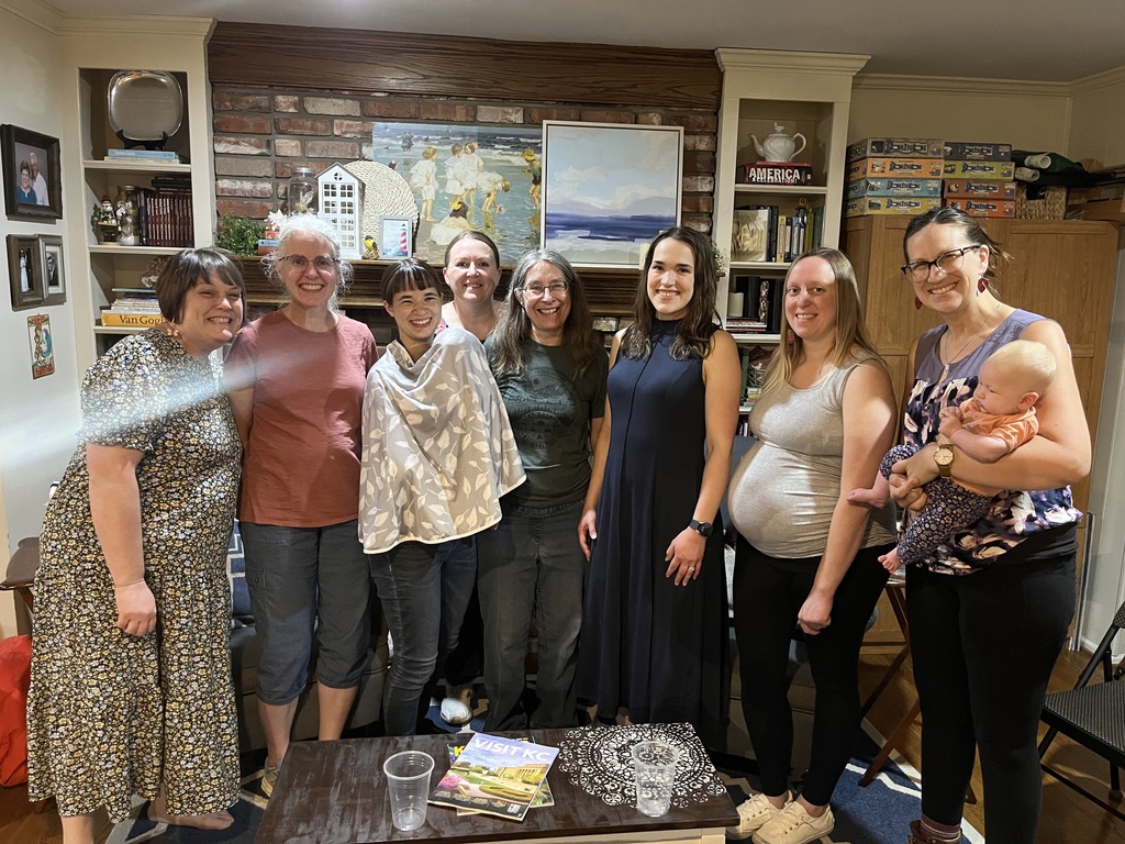 Group of eight women and one child standing indoors in front of a brick wall with paintings and shelves.