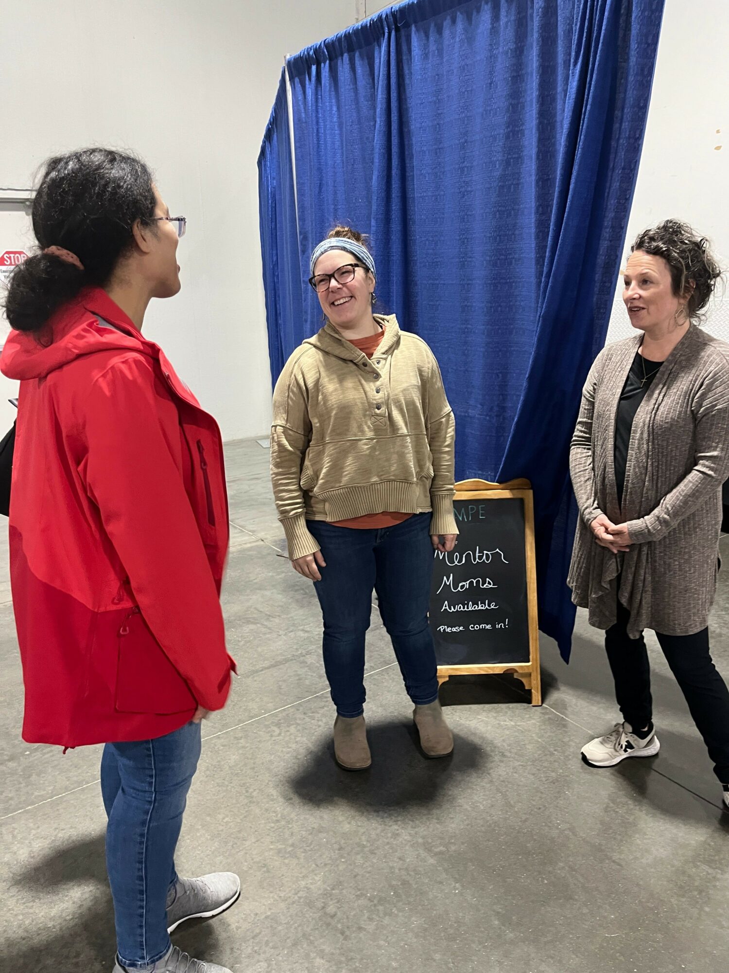 Two women standing and talking indoors, with a blue curtain and a chalkboard in the background.