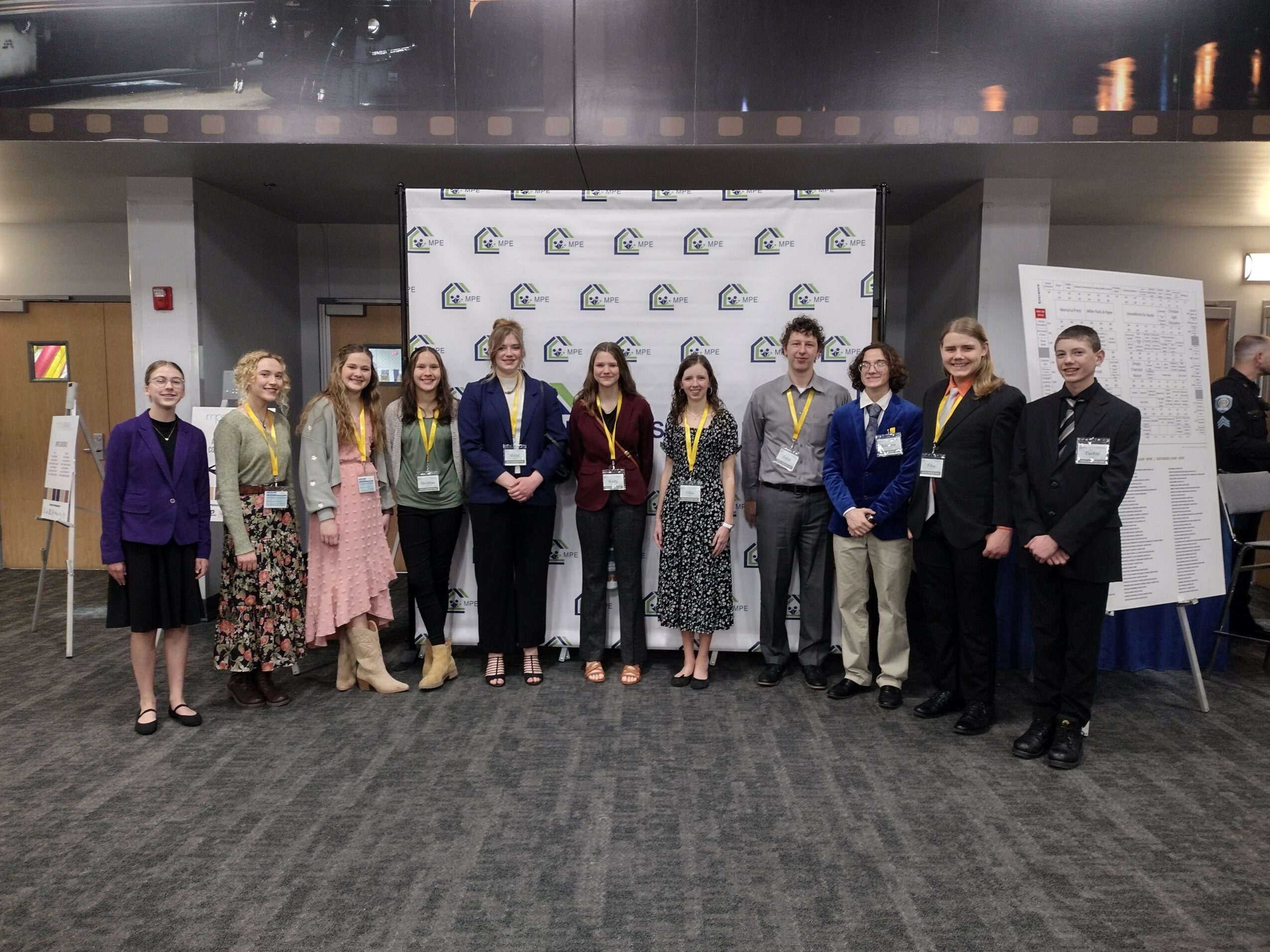 Group of eleven people standing in a line at a conference, with a backdrop and presentation board behind them.